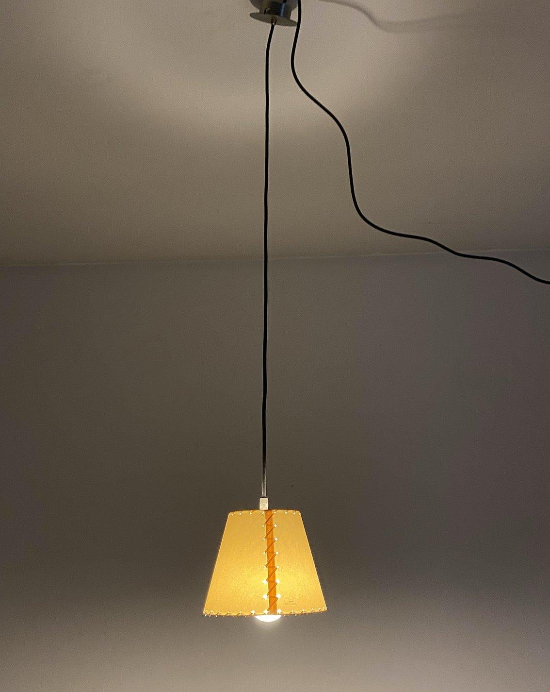 Lampada a sospensione SiSiSi prodotta da Santa e Cole in Spagna nel 1990. Con una struttura in metallo, la lampada si caratterizza per un paralume a forma conica in pergamena, arricchita da una cucitura a mano in tessuto. La preziosità dei suoi