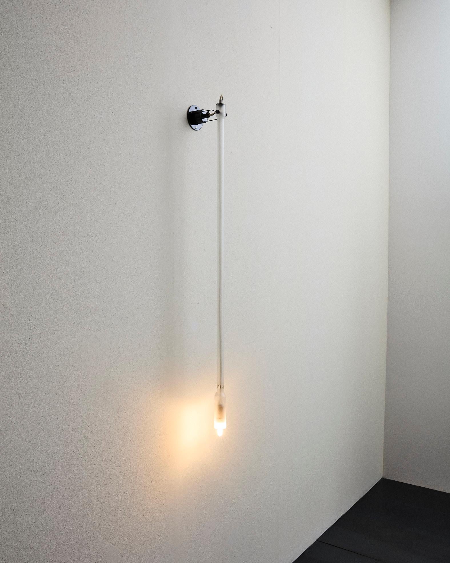 Lampada orientabile modello “2702 Canna nuda” composta da una struttura in metallo e una canna in vetro satinato che alloggia la sorgente luminosa. Il corpo può essere reclinato di 90°, consentendo l’utilizzo sia a parete sia a