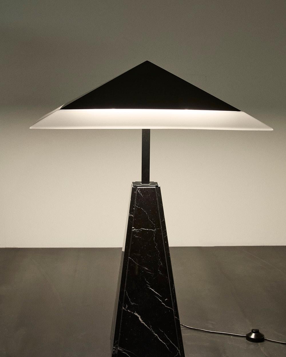 Elegante e scultora, la lampada da tavolo 'Abat Jour', disegnata da Cini Boeri per Arteluce nel 1975 è un'icona del design italiano.
La base piramidale in marmo nero conferisce solidità e prestigio, mentre il diffusore a forma di piramide tronca, in