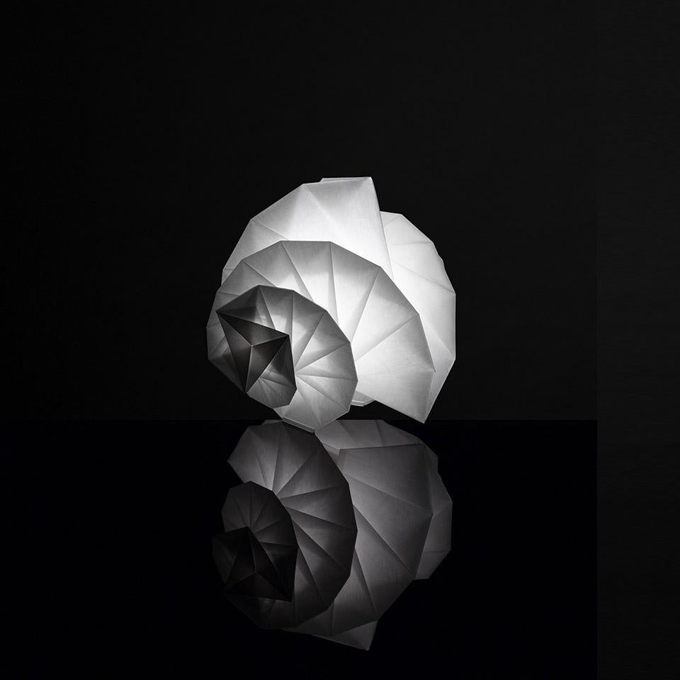 Lampe de table modèle Mendori IN-EI du fabricant Artemide en collaboration avec Reality Lab et conçue par Issey Miyake
La lampe est fabriquée à partir de matériaux recyclés qui lui confèrent une transparence lumineuse supérieure à celle du papier.
À