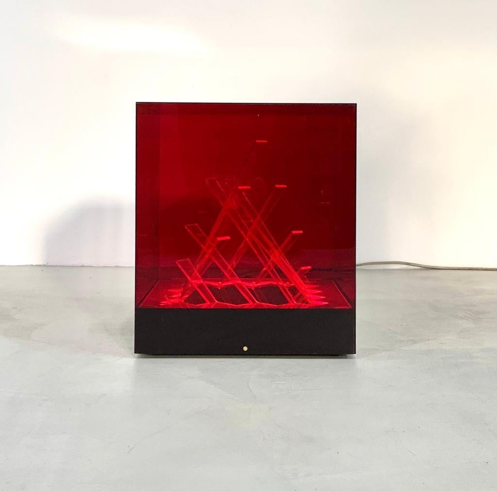 Ampada da tavolo mod. Cubo di Teo in vetro acrilico rosso.
Questa scultura cubica presenta al suo interno una serie di rettangoli curvati e intrecciati tra loro. 
Al di sotto è presente la fonte luminosa che permette di creare un gioco di riflessi