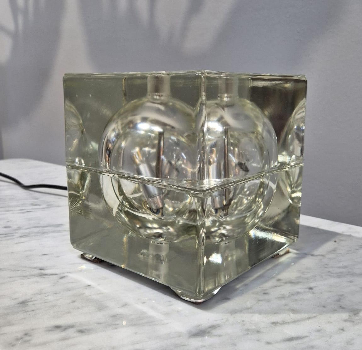 Tischleuchte Cubosfera, entworfen von Alessandro Mendini im Jahr 1968.
Diese von Fidenza Vetraria hergestellte Lampe besteht aus kristallinem Glas, das zu einem Würfel geformt ist. Die Leuchte steht auf vier Metallbeinen und ist mit zwei