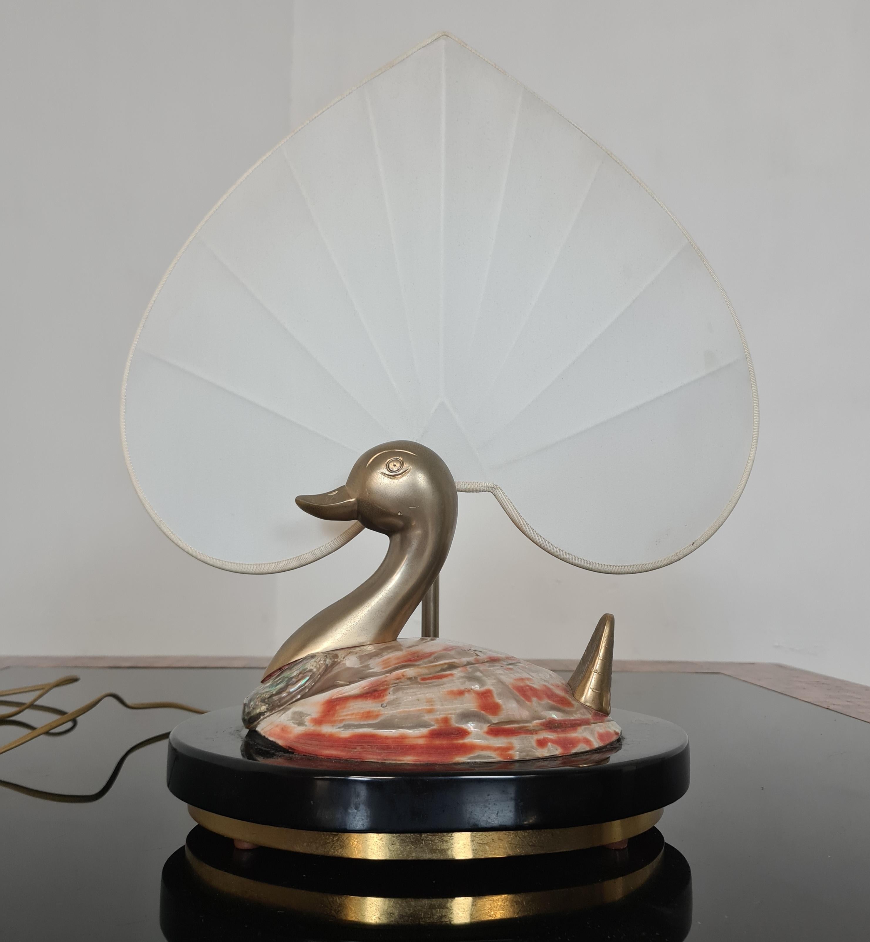 Table lamp by Antonio Pavia.

Refined 80' Hollywood Regency style table lamp

The lamp features a black base with brass profiles on which there is a duck with a shell body and brass head and tail.

Complementing it is a large fabric lampshade in the