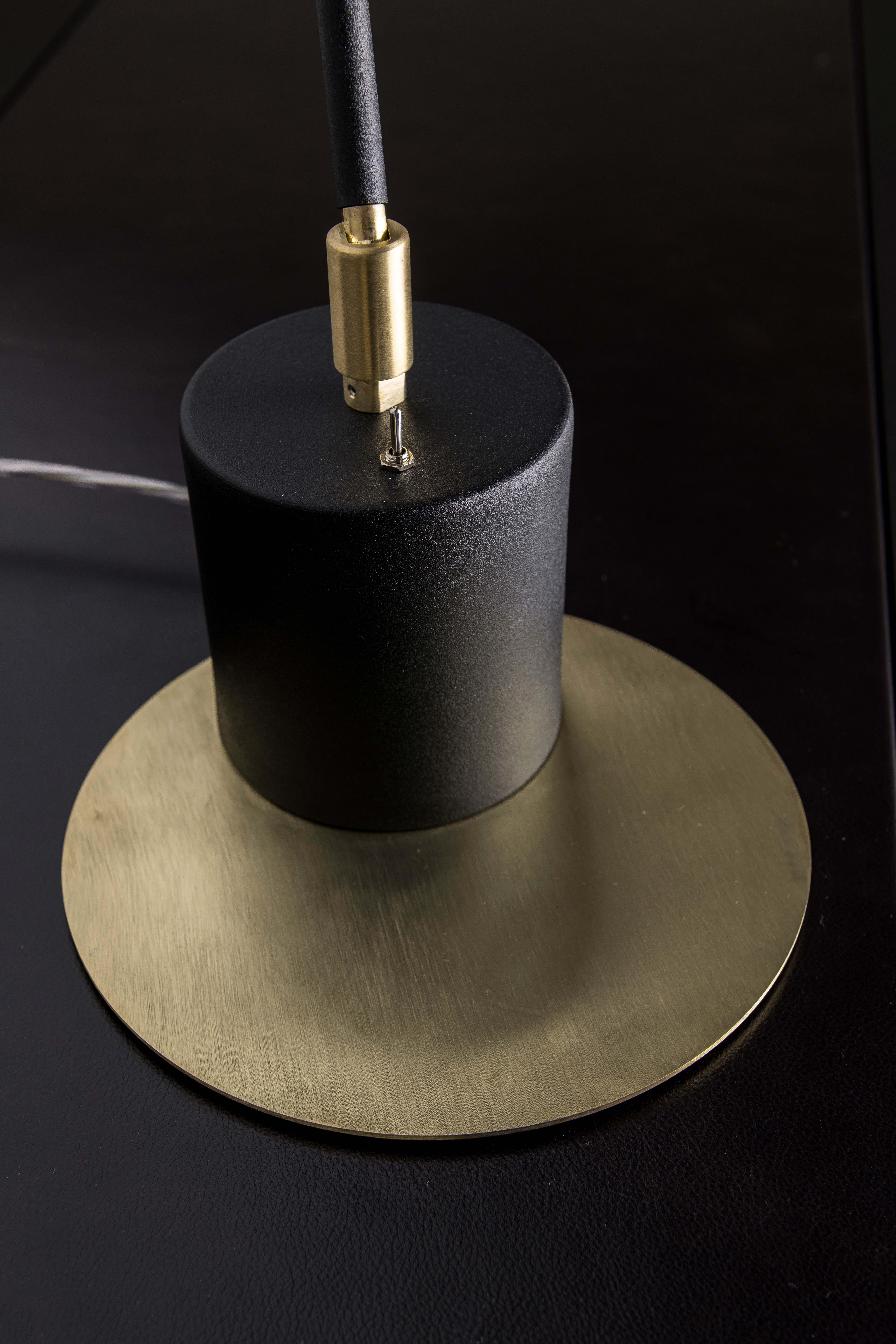 Table lamp with an essential and modern line, embellished with natural satin brass details. Table lamp with matte black powder-coated aluminum frame and lamp body with circular satin brass plate. The joints are made of brushed brass. It can also be