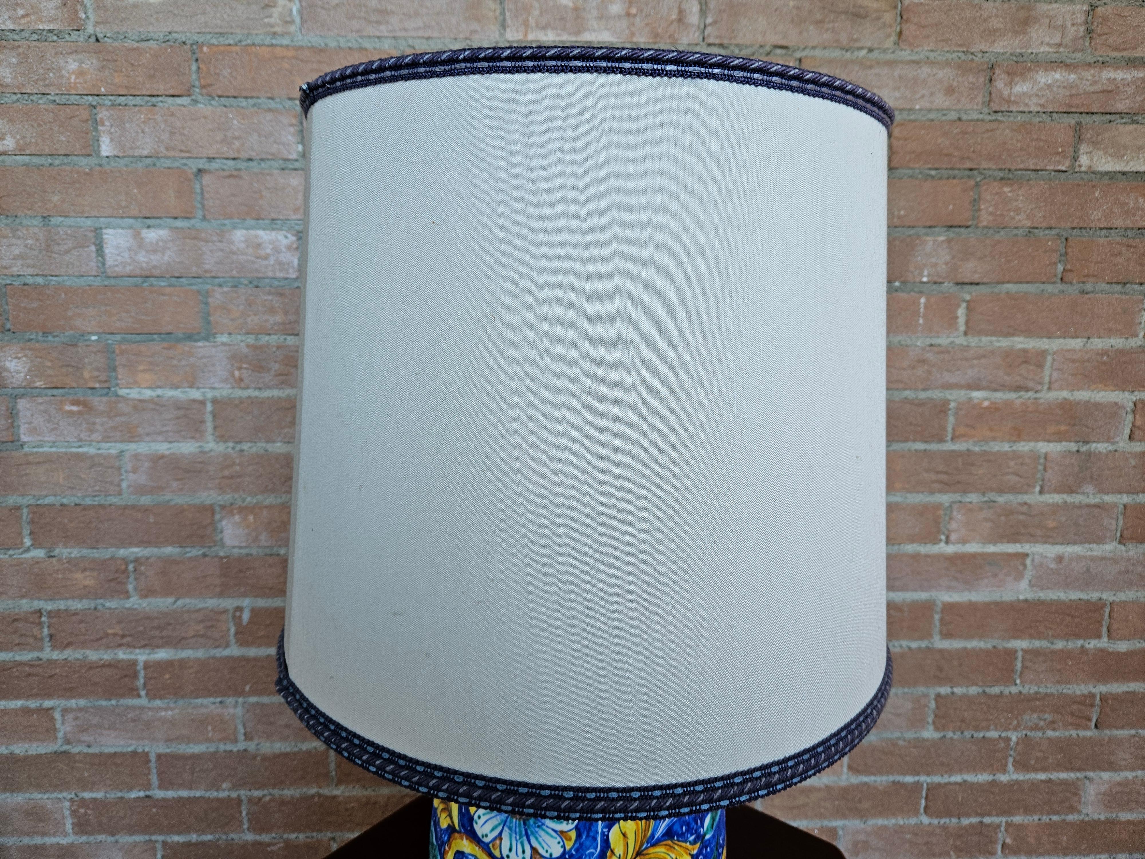 Bunt und lebendig bemalte Keramik-Tischlampe mit cremefarbenem Stoffschirm und blauen Verzierungen.

Glühbirne nicht enthalten.
