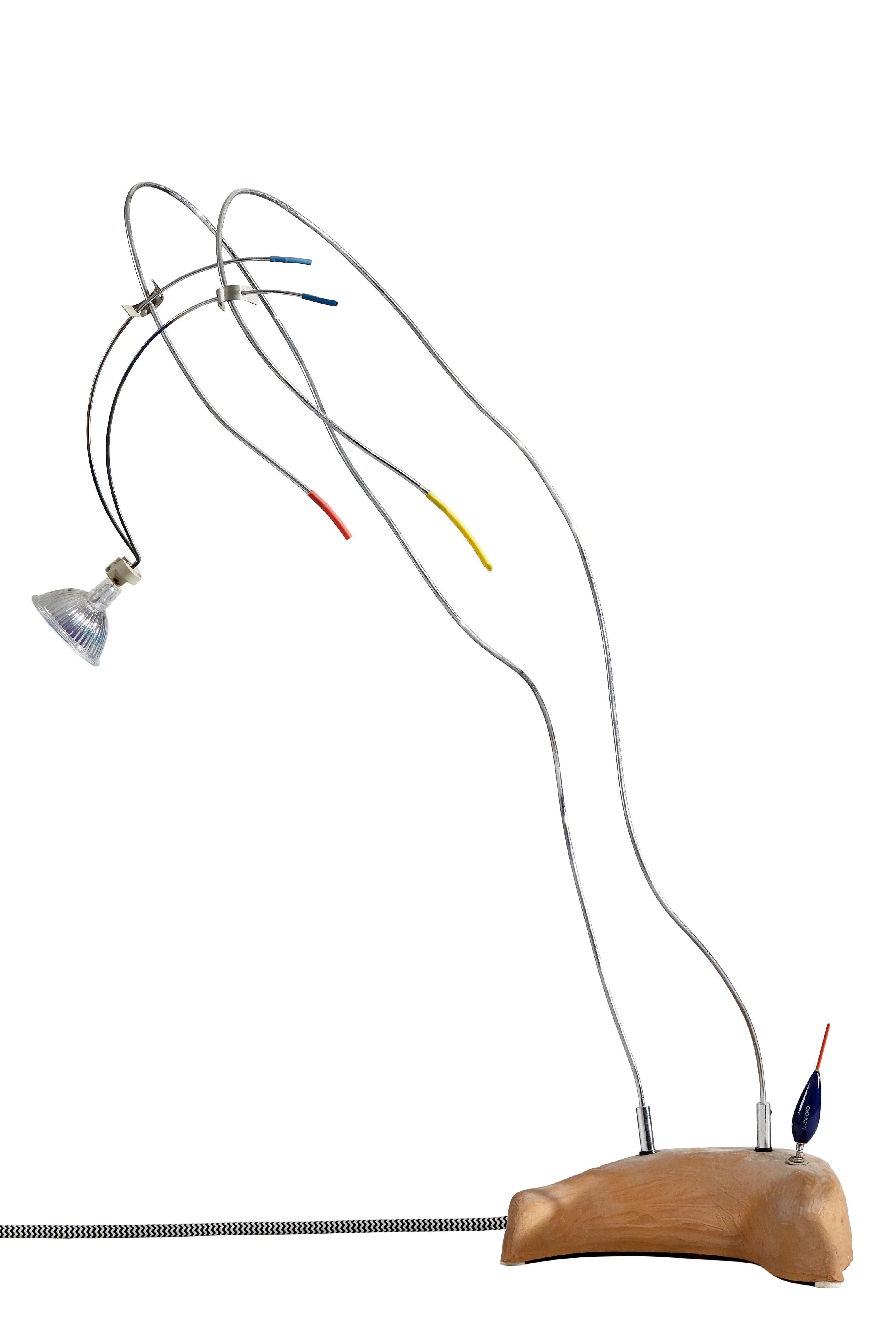Lampada da tavolo Titivilus disegnata e prodotta da Lucifero Design. Il prodotto si compone di una struttura sinuosa e ondulata, caratterizzata da cavi a vista che collegano la fonte luminosa al corpo centrale tramite fili sottili in acciaio
