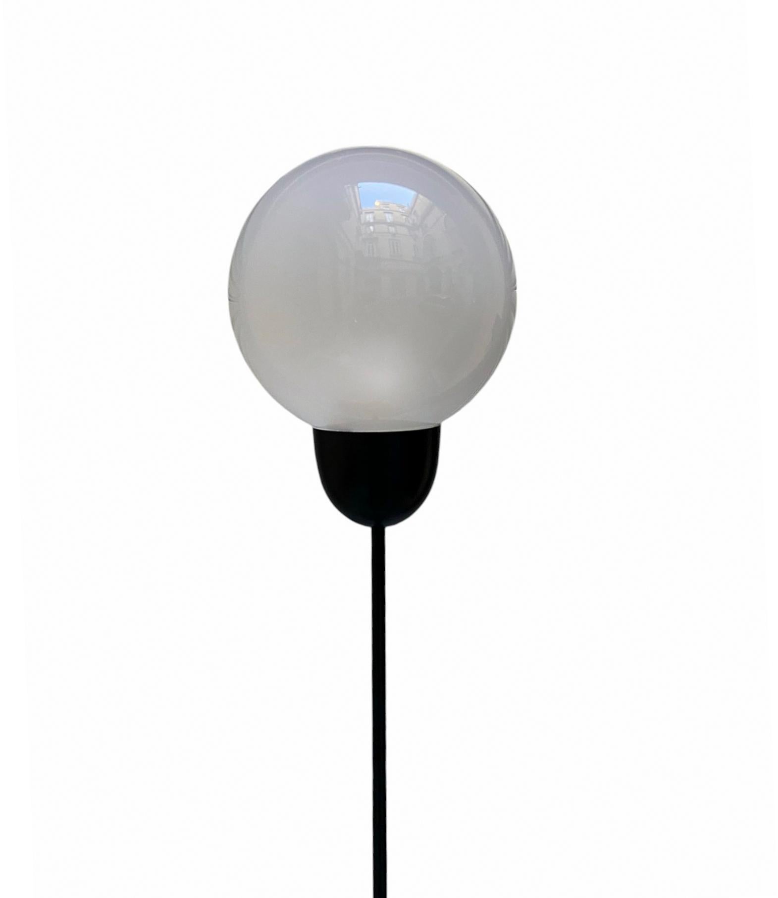 Lampadaire des années 1970  caractérisé par une tige verticale élancée s'élevant à partir d'une base circulaire. La tige est en métal peint en noir. À l'extrémité supérieure de la tige, un diffuseur sphérique en verre opale blanc est positionné et