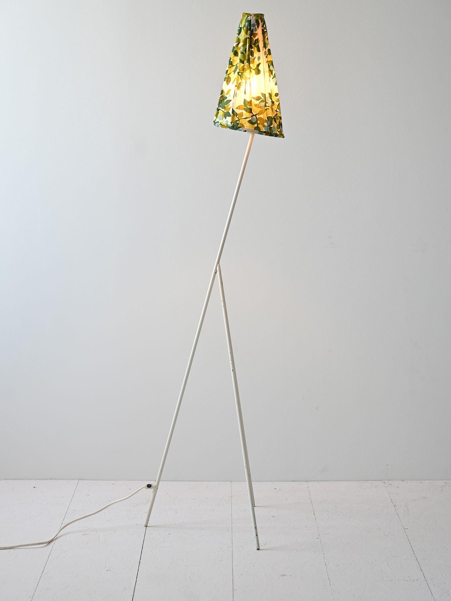 Vintage lamp characterized by a white tubular metal stem, tilted forward and supported by three feet.
The cylindrical fabric shade is decorated with leaf motifs, introducing an element of naturalness and artistic detail. These delicate and stylized