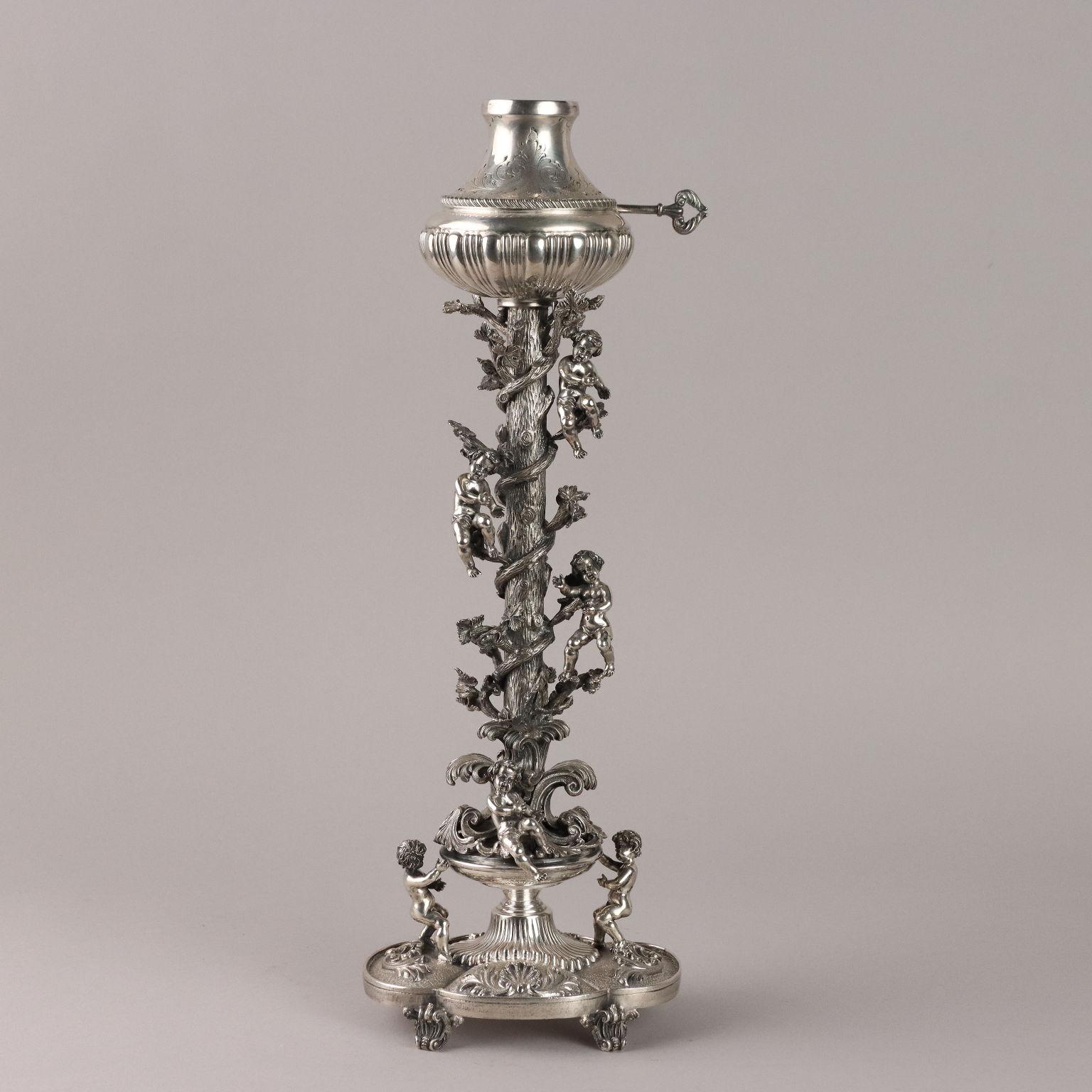 Lampada in argento sbalzato, baccellato e finemente cesellato. Fusto raffigurante tronco d'albero cinto da tralci di vite e putti. Sotto la base marchi dell'argento incussi, firma dell'argentiere incisa e etichetta del rivenditore applicata. Grammi