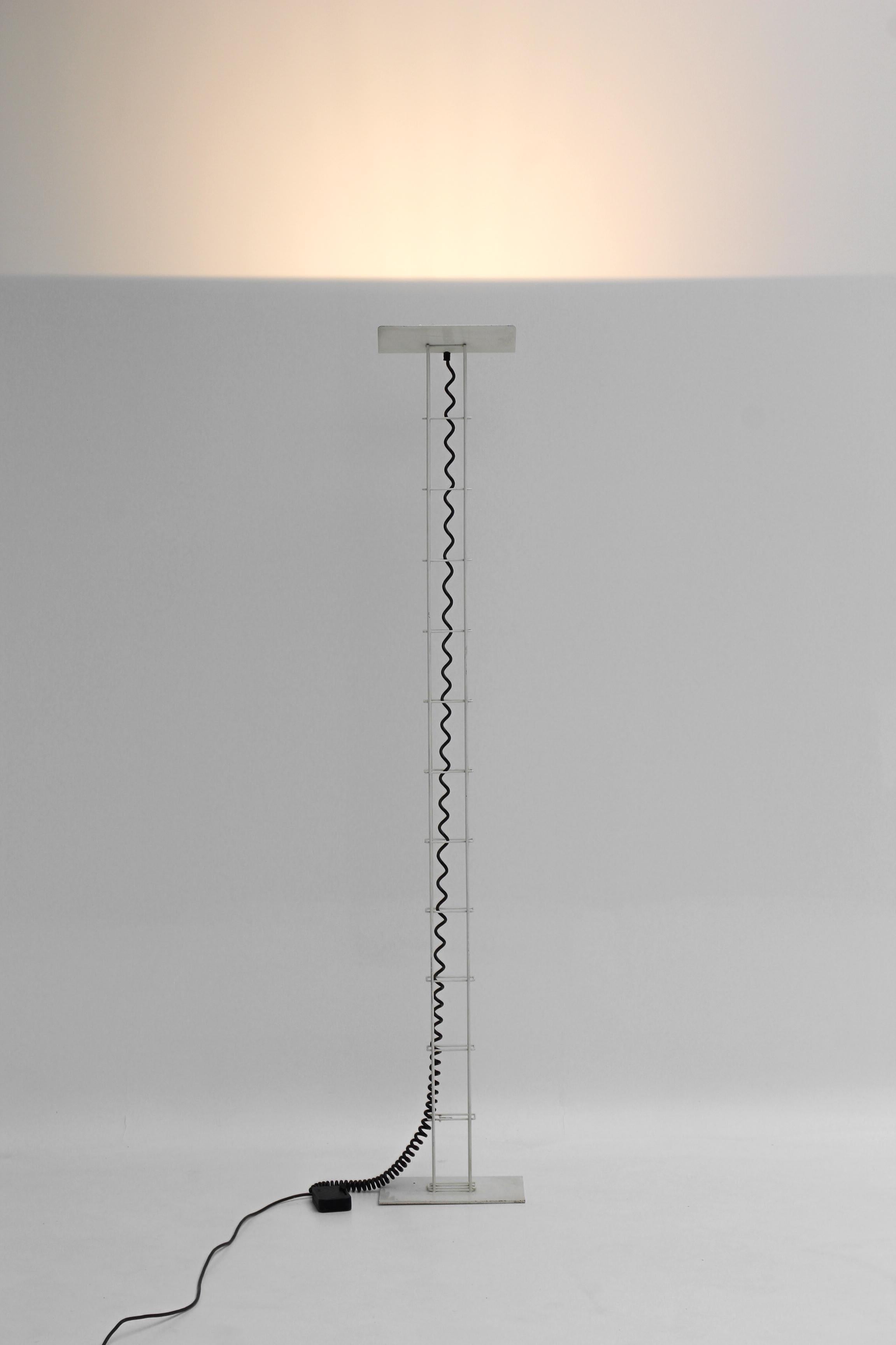 Édité par Arelux en Italie dans les années 1980, ce lampadaire minimaliste séduit par sa silhouette élancée et sa lumière indirecte.

Sa structure tubulaire en métal laqué blanc repose sur une base fine et stable, tandis que le câble spiralé
