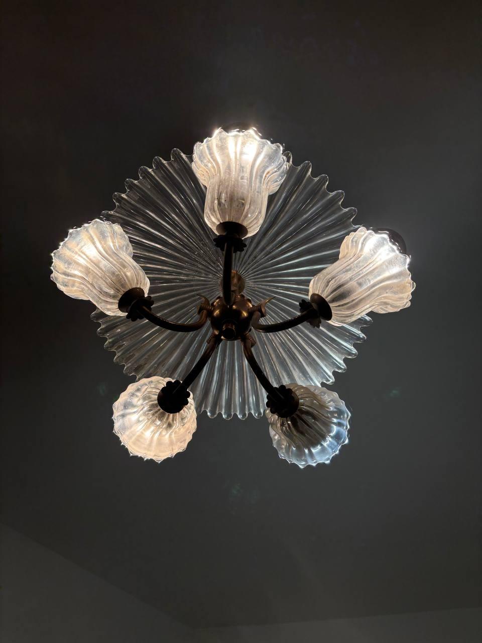 Cet exquis lustre Art déco en verre de Murano, créé par le célèbre designer Ercole Barovier, incarne l'élégance et le savoir-faire vénitiens des années 1930 et 1940. Sa structure centrale en métal supporte un auvent supérieur décoré et un élément