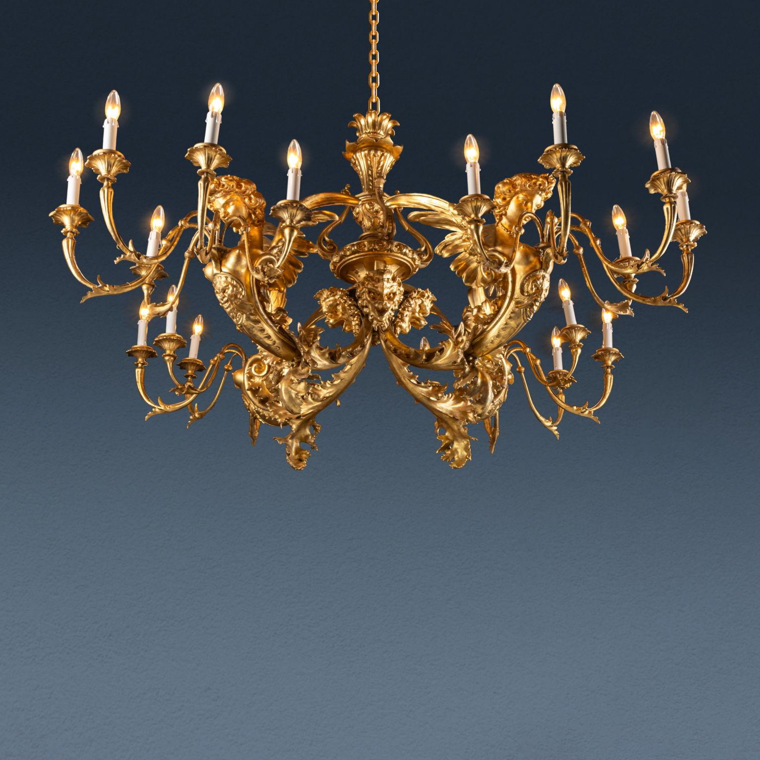 Carved and gilded metal and wood chandelier. The central body, carved with floral and leaf motifs, is ornamented at the bottom with four lion heads, alternating with as many arms carved with curling leaf volutes and ending with busts of winged