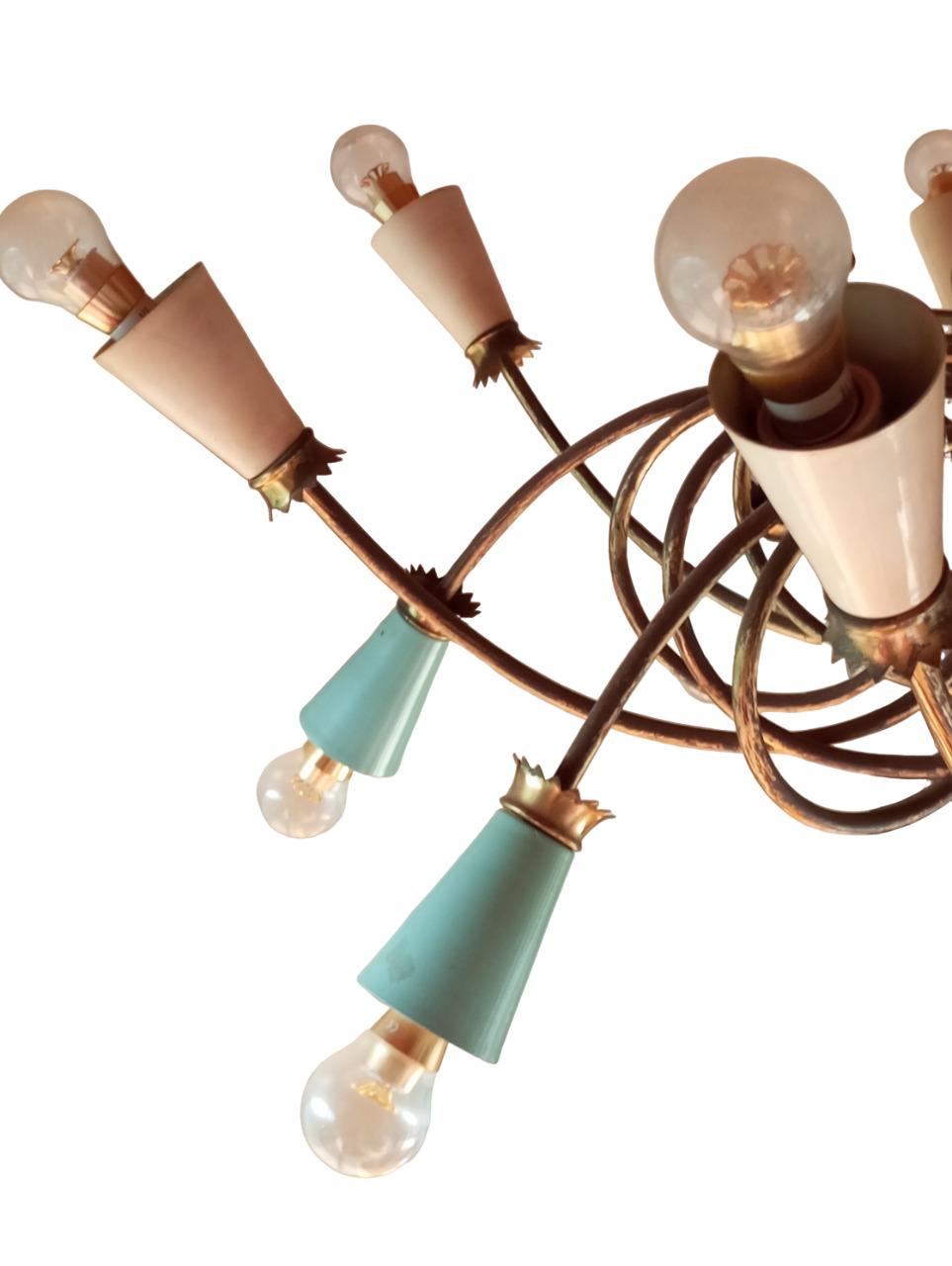 Il lampadario descritto è un elegante pezzo vintage degli anni '50, ispirato allo stile dei grandi designer dell'epoca come Angelo Lelli, Pietro Chiesa, Tommaso Buzzi, Fontana Arte e Giò Ponti. Realizzato in ottone, presenta una struttura raffinata