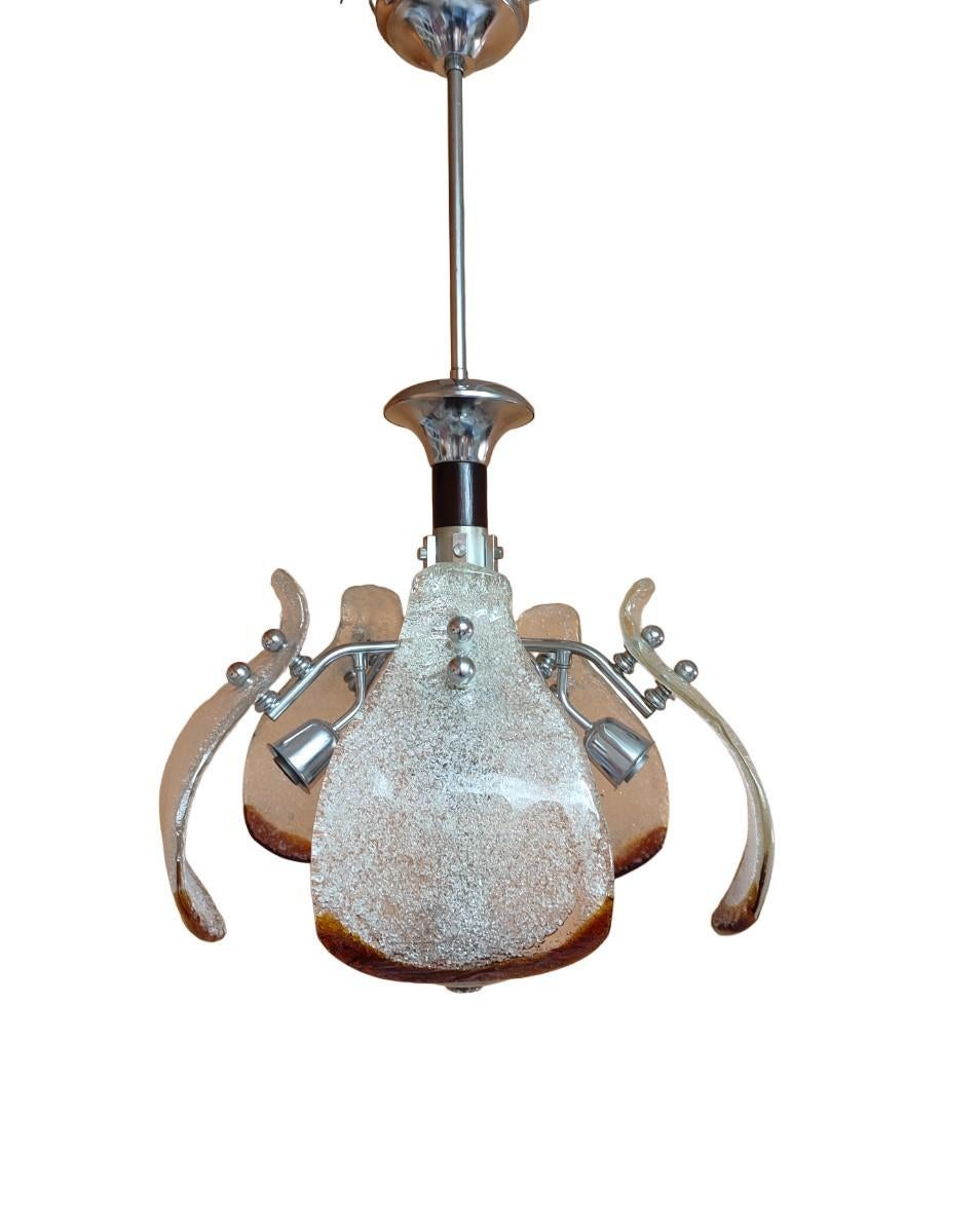 Esta lámpara de araña es un fascinante ejemplo del arte del cristal de Murano, producido en la década de 1970 por la renombrada empresa Mazzega, y combina tradición y modernidad a través de una estructura de metal cromado.
La estructura metálica
