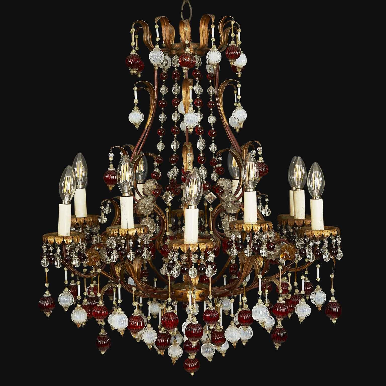 Twelve-light chandelier  on two tiers of height, made in Italy in about 1960. This chandelier has a gilded iron cage frame, circular in shape, twelve curved arms adorned with foliate elements and richly decorated with colored glass, chains of red
