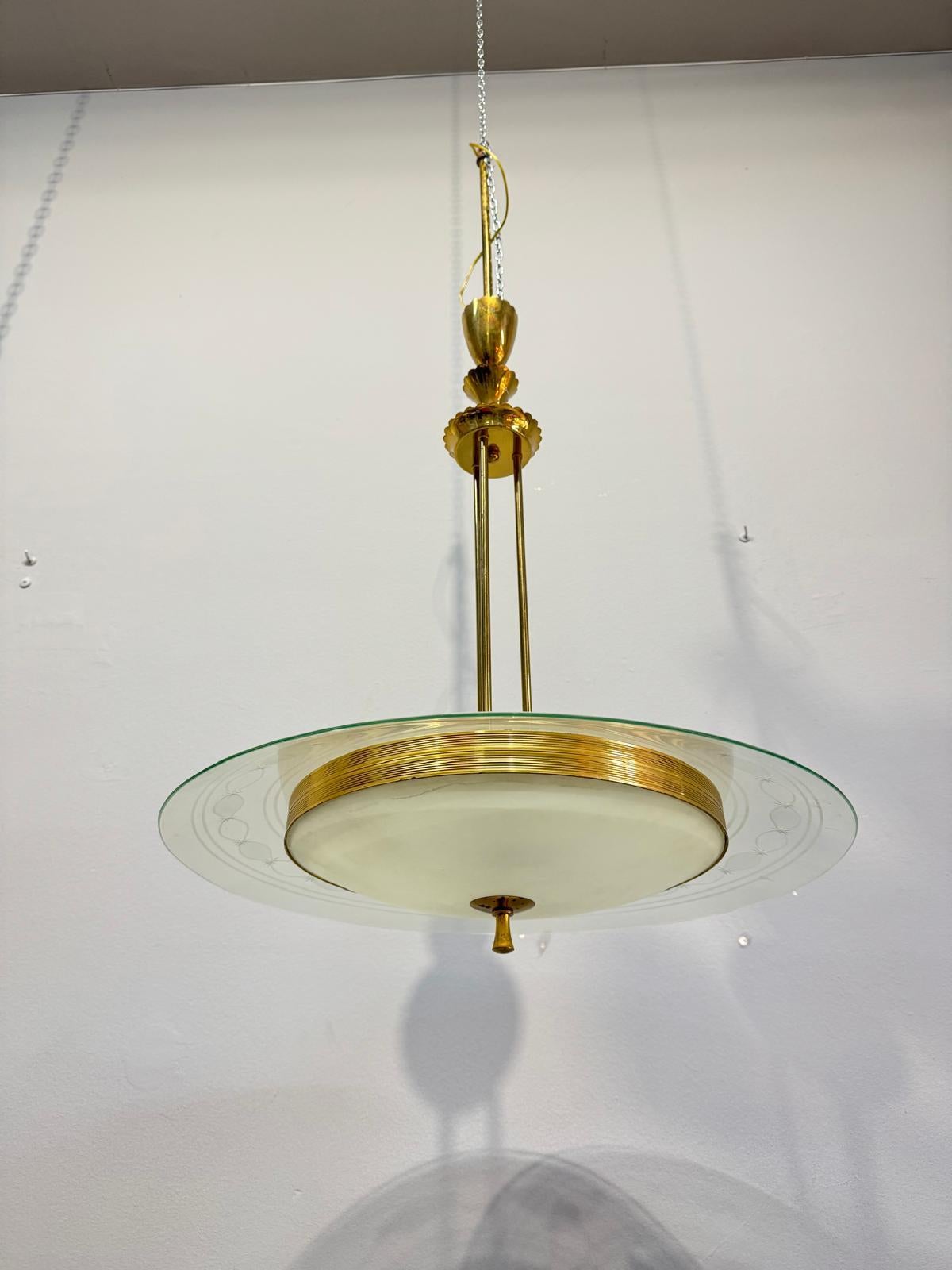 Lampada a sospensione disegnata da Pietro Chiesa. Il lampadario presenta una struttura in ottone con un vetro principale incolore con un motivo disegnato e un vetro opalino più piccolo, sorretto da un disco in ottone.

