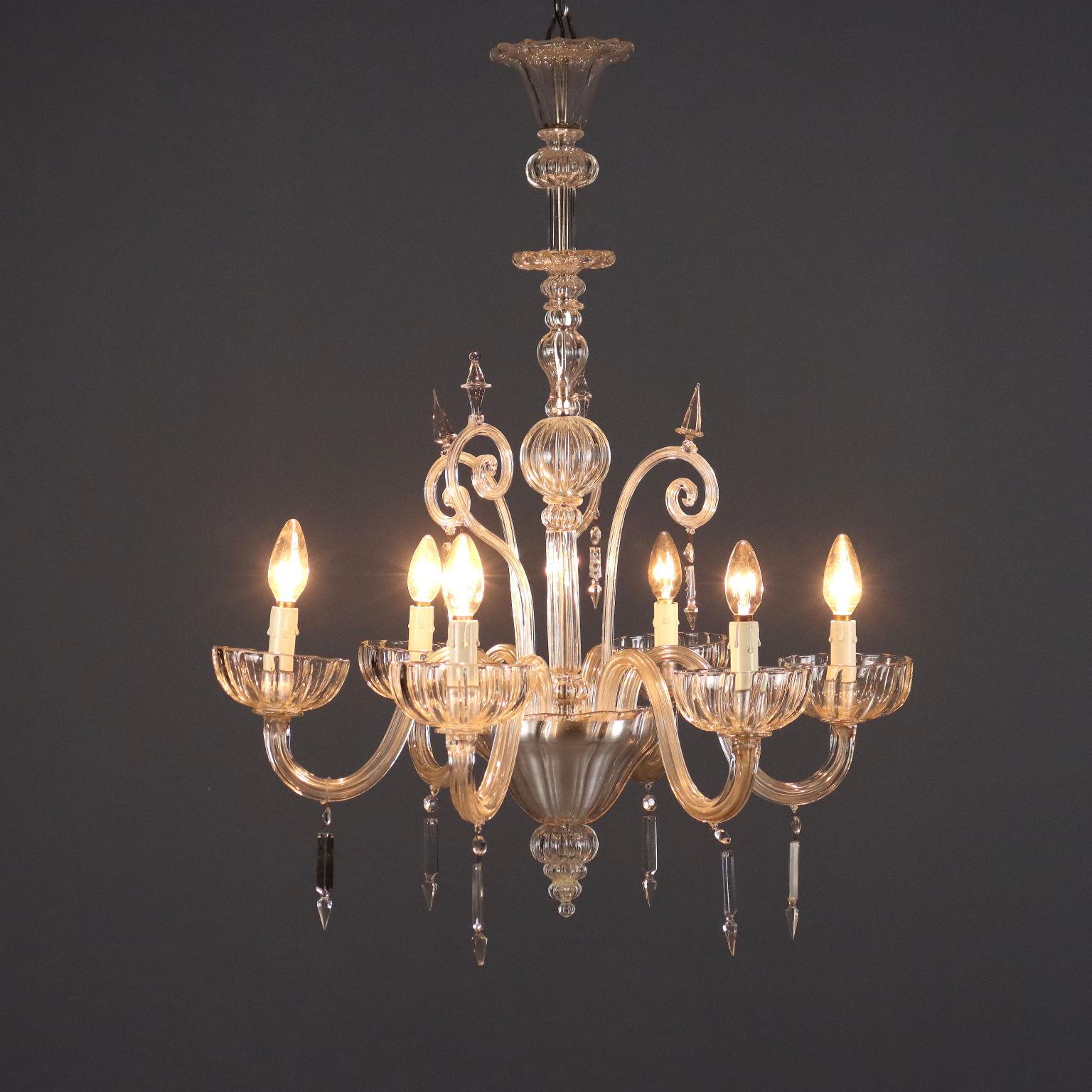 Lampadario Veneziano in vetro soffiato trasparente. Sei luci sorrette da braccia mosse, fusto centrale arricchito da gruppo di elementi a ricciolo di cui due mancanti.
Murano, fine XIX Secolo.	
