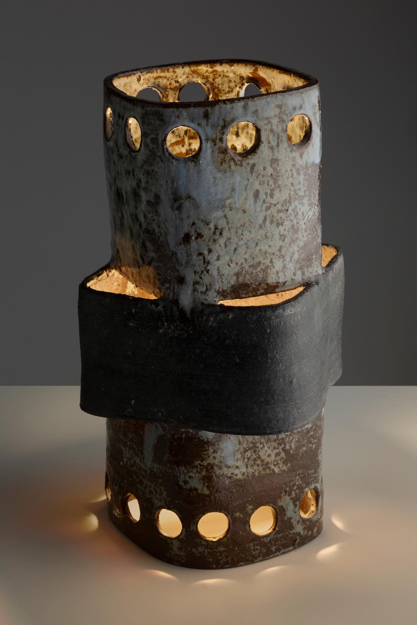 Esta lámpara escultural de Cécile Dein destaca por su forma arquitectónica y su materialidad bruta. Fabricada en cerámica, tiene un cuerpo cilíndrico salpicado de perforaciones circulares que difunden una luz cálida y ambiental. Una audaz banda