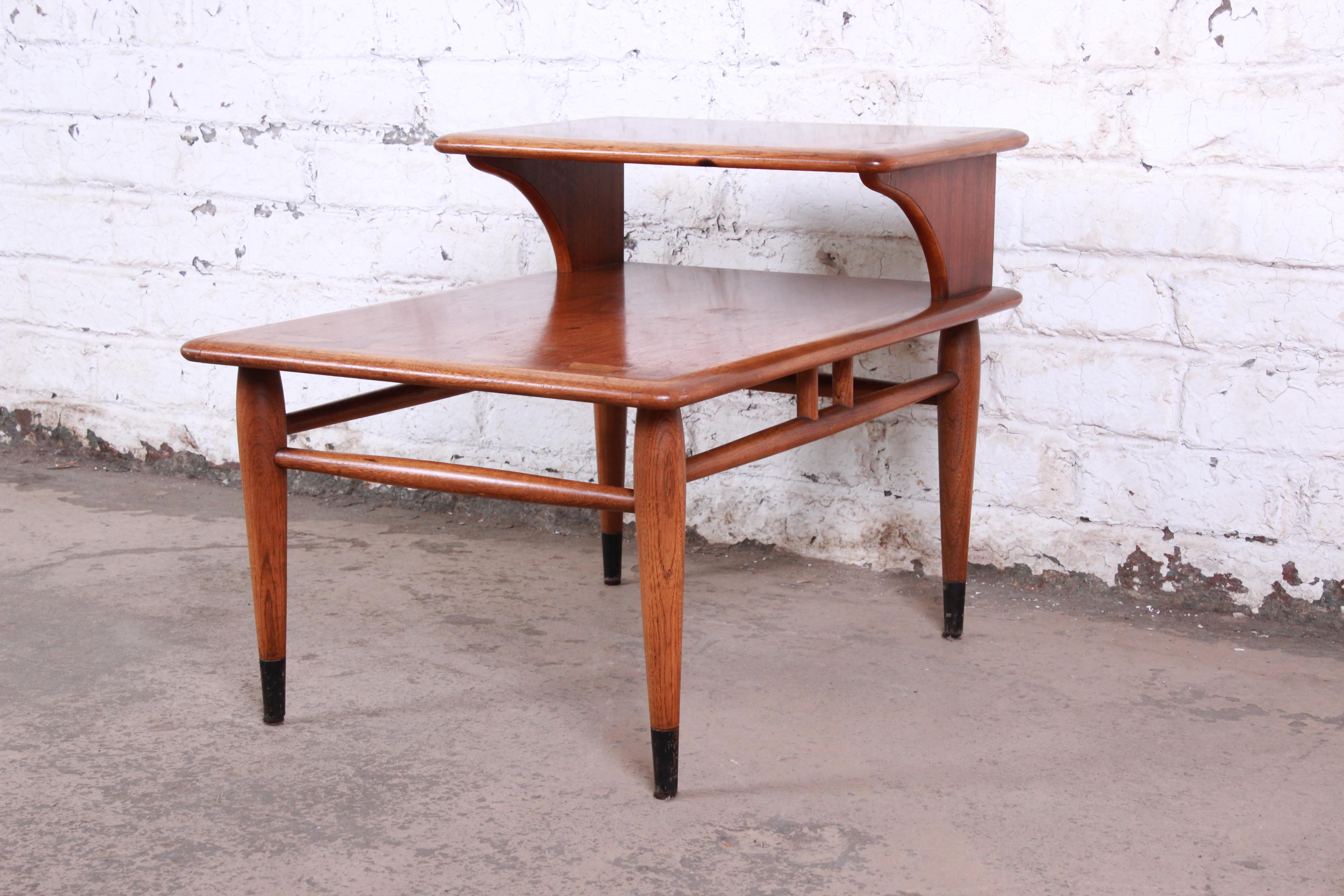 A gorgeous Mid-Century Modern step end table from the Acclaim line by Lane. The table features beautiful walnut and ash wood grain and a unique dovetail design. It sits on tapered legs with ebonized feet and sleek stretchers connecting the legs. The