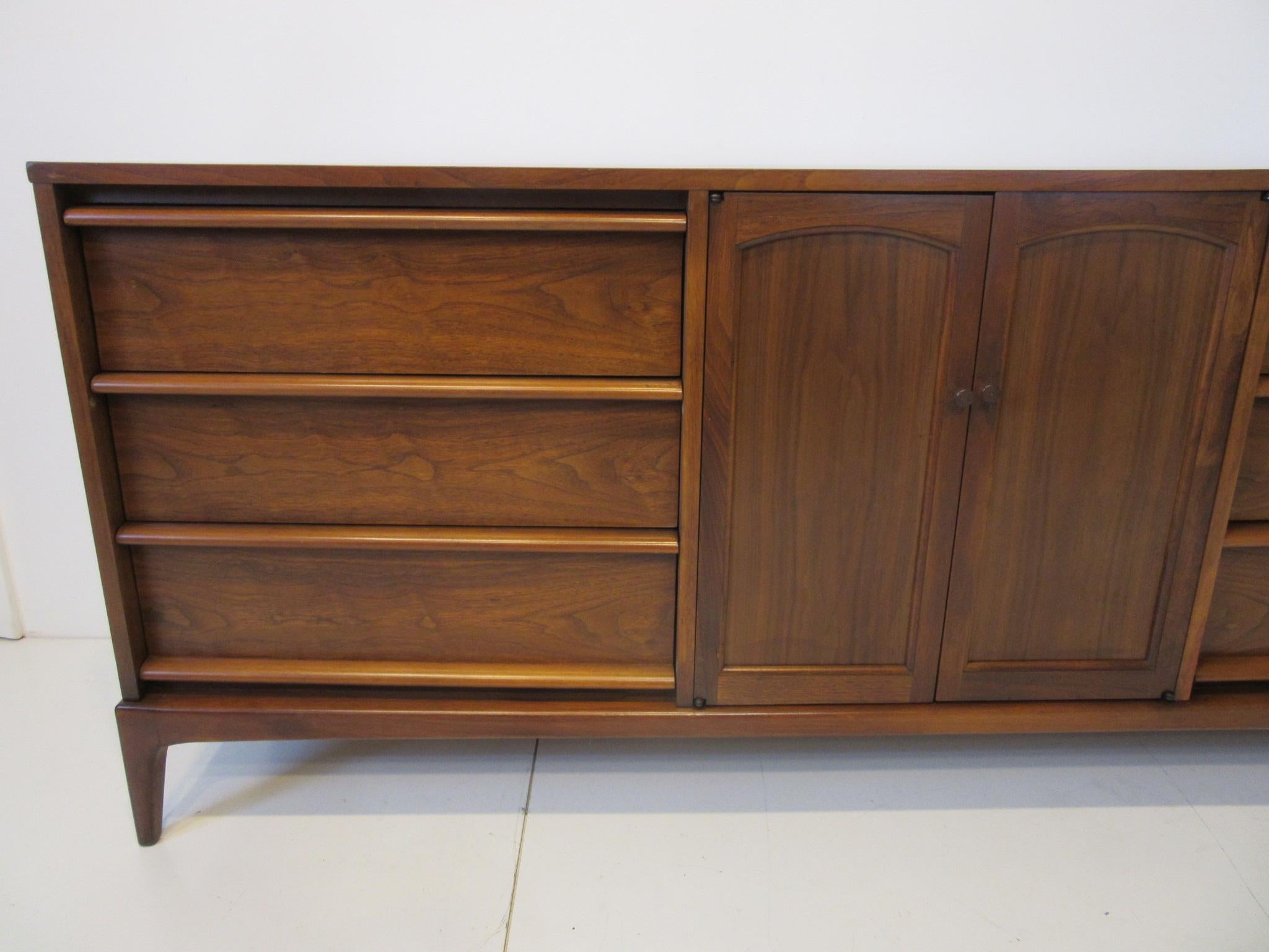 A walnut midcentury minimalist dresser with six drawers and two doors revealing three additional drawers. The doors have reversible panels in matching walnut or the other side having woven caning all sitting on beautifully crafted slender legs for a