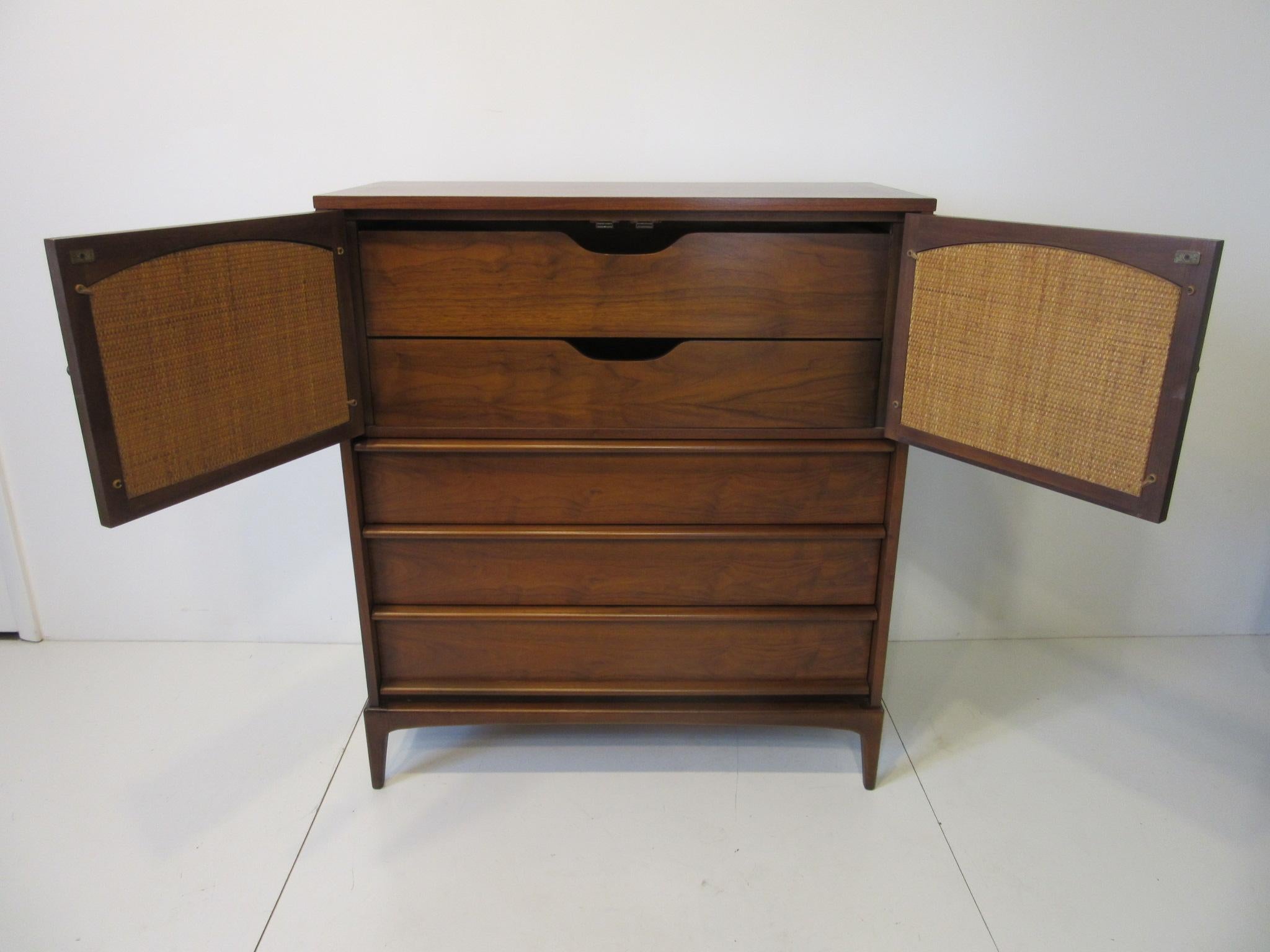 A tall dresser chest in a dark walnut with plenty of storage with lower drawers and upper doors opening to reveal additional storage. The doors have reversible panels with matching walnut or woven caned fronts and having slender legs giving the