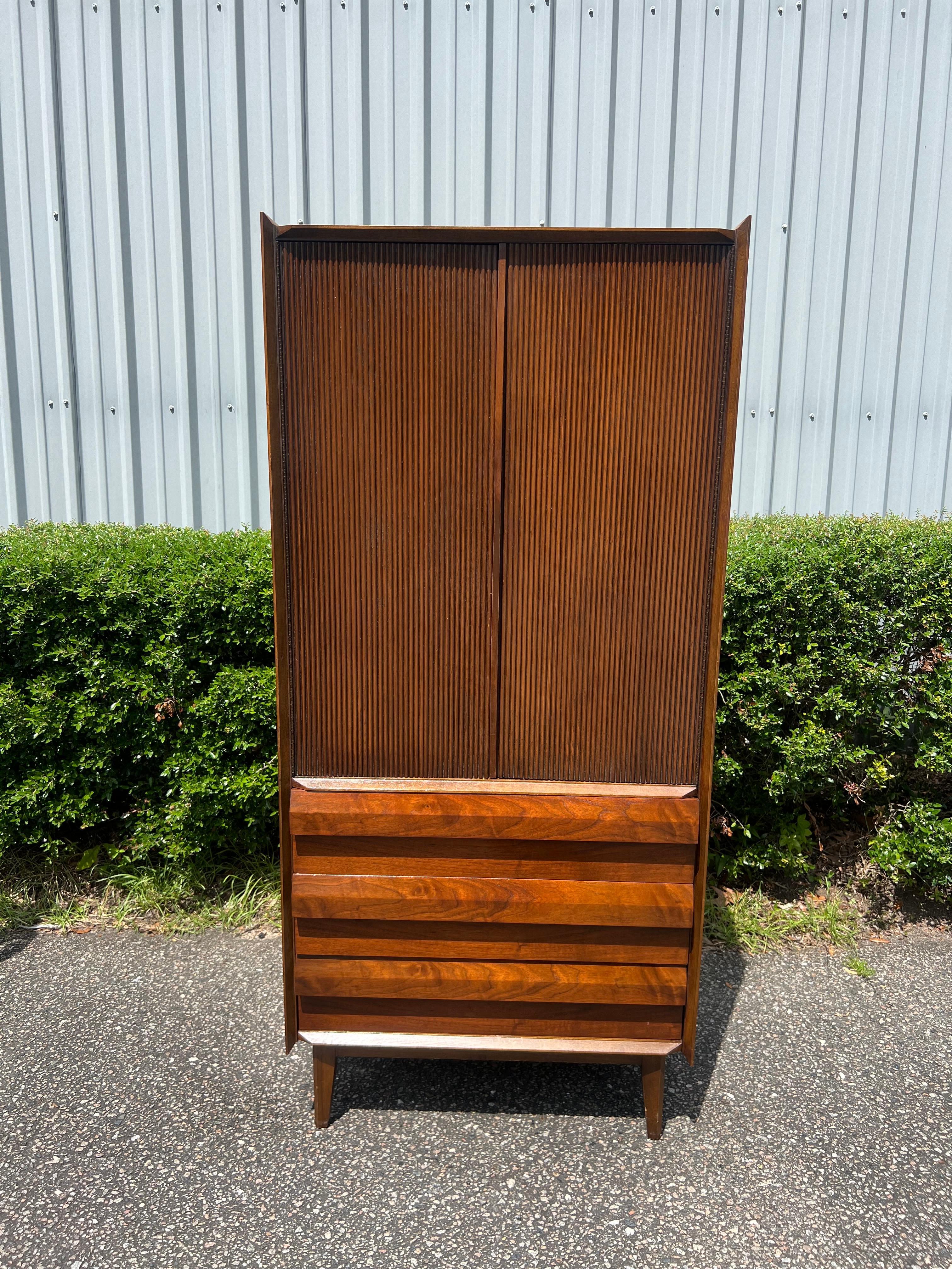 This Beautiful and sturdy walnut mid century modern tall gentleman’s armoire from Lane has a great masculine and elegant presence.
The ribbed doors contain 1 pullout drawer with shelving above. When the doors are open, you have a mirror on one side