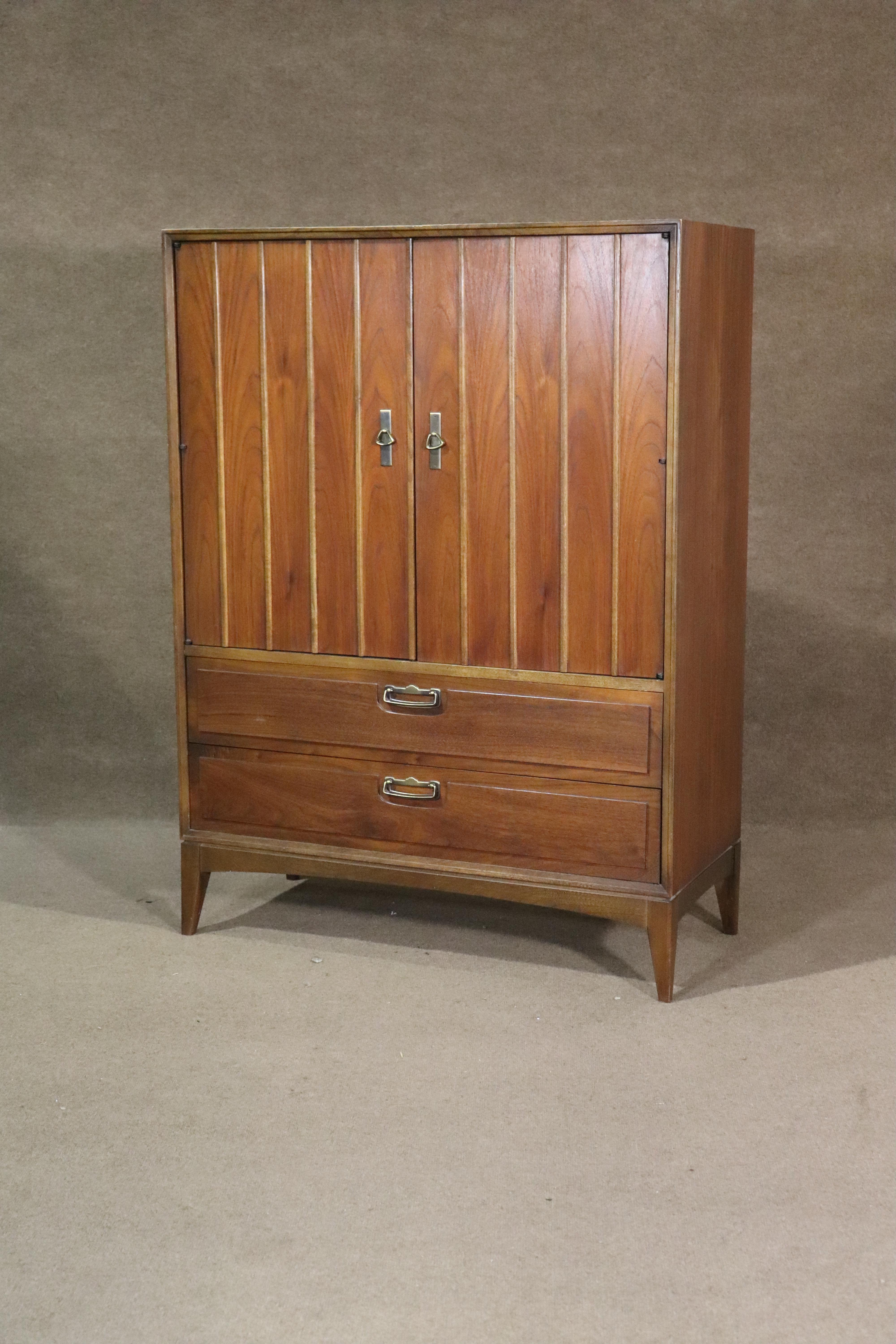 Cómoda alta de Lane Furniture en madera de nogal veteada. Esta cómoda de caballero tiene cuatro amplios cajones y almacenamiento abierto. Dos grandes puertas con herrajes de latón y patas cónicas. Un atractivo complemento de mediados de siglo para