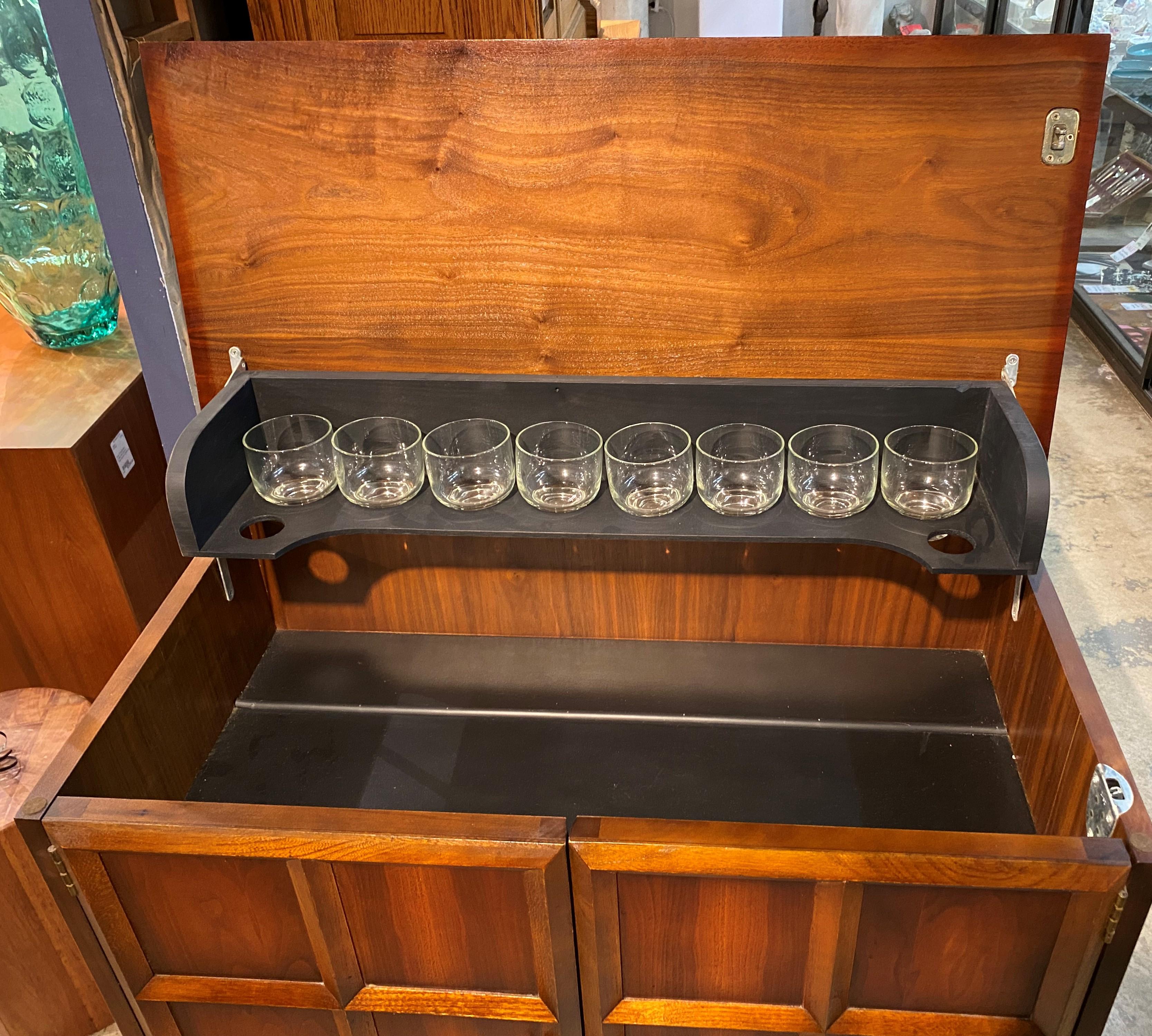 A fine midcentury walnut two door bar or liquor cabinet with lift top by Lane Furniture, opening to glassware storage on the hinged top, as well as on each door, with eight larger glasses on each door, and eight smaller glasses on the top rack. The