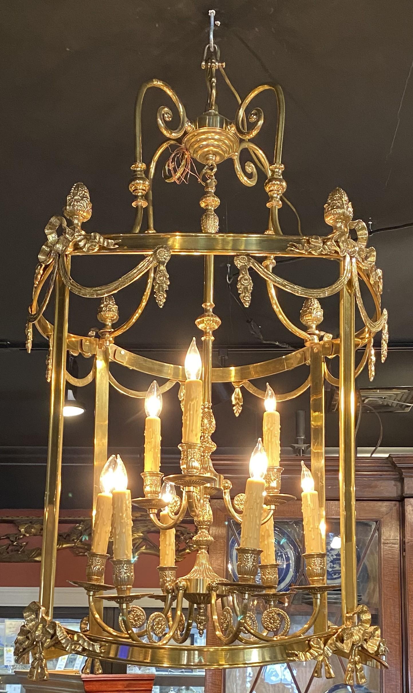 Un meraviglioso lampadario a nove luci elettrificate a forma di lanterna in ottone con bracci a voluta, decorazione a nastro e rosette in ottone. Risale al XX secolo ed è in buone condizioni generali di funzionamento, con lievi imperfezioni