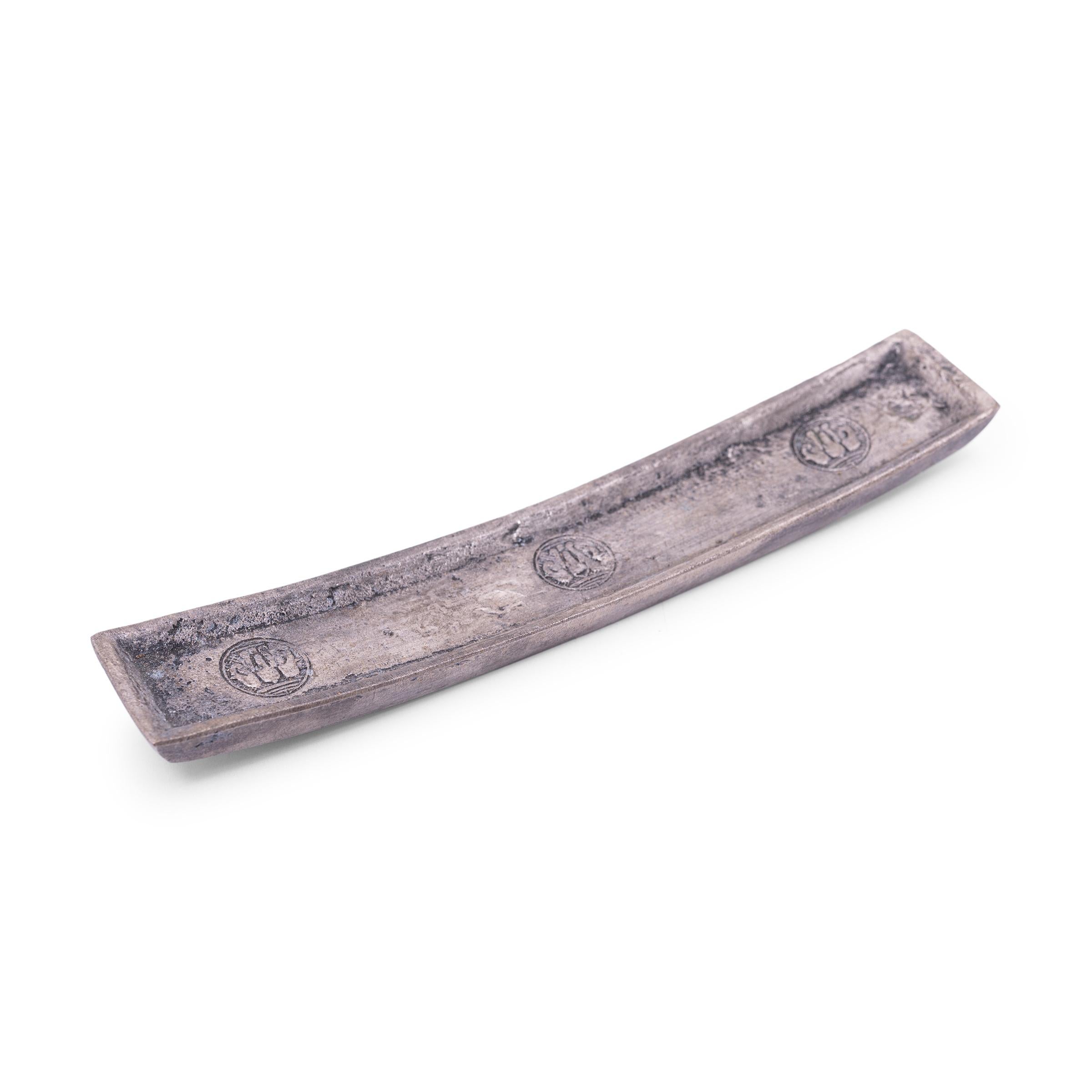 This silver alloy ingot is cast in the form of an elongated bar, with a slender, rectangular shape and curved ends. Resembling a shallow dish or incense stand, the ingot has a concave surface stamped with three round medallions of a three-headed