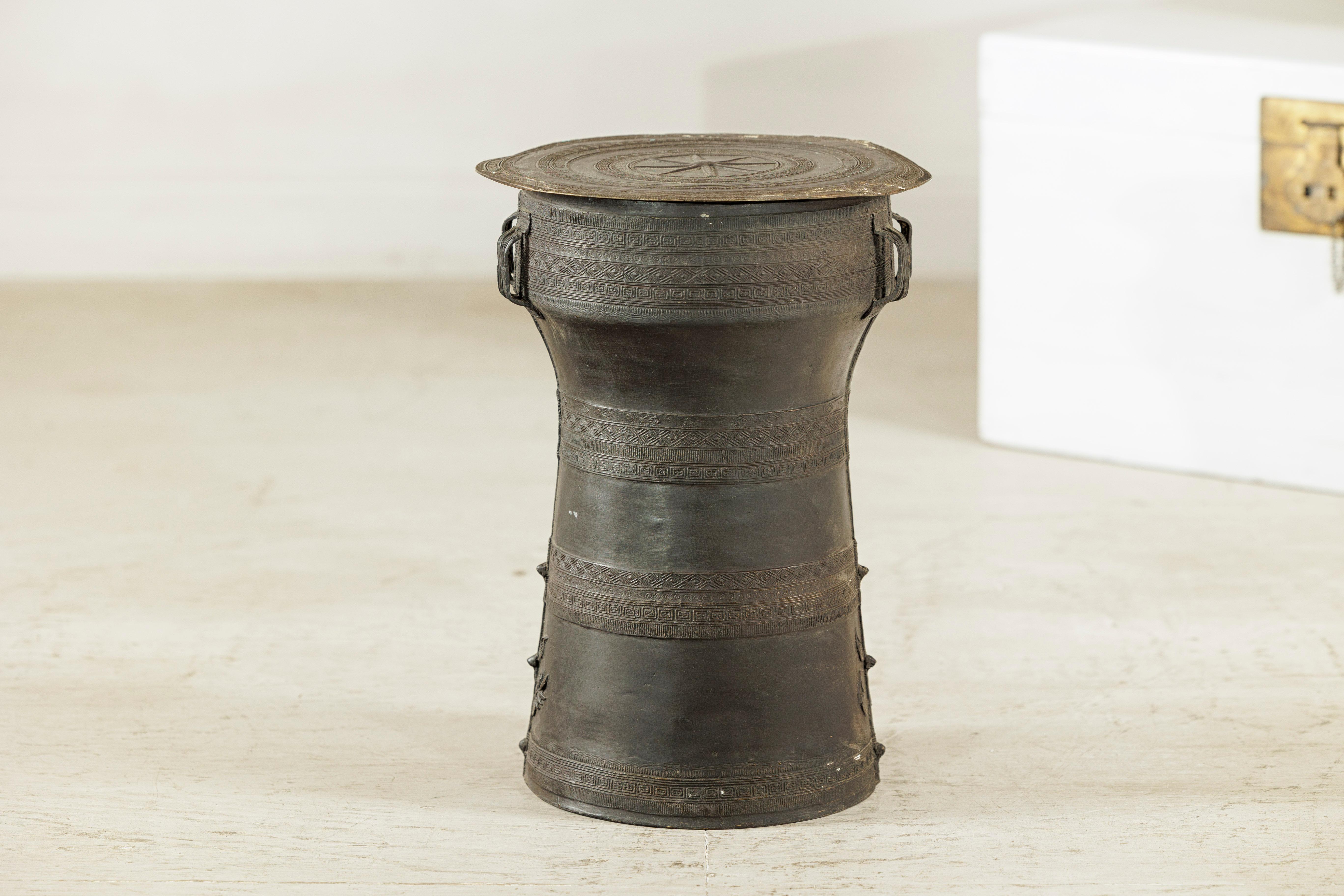 Este tambor de lluvia de bronce laosiano presenta una característica forma de reloj de arena y una pátina oscura envejecida de forma natural. La parte superior circular está decorada con un medallón en forma de estrella rodeado de bandas geométricas
