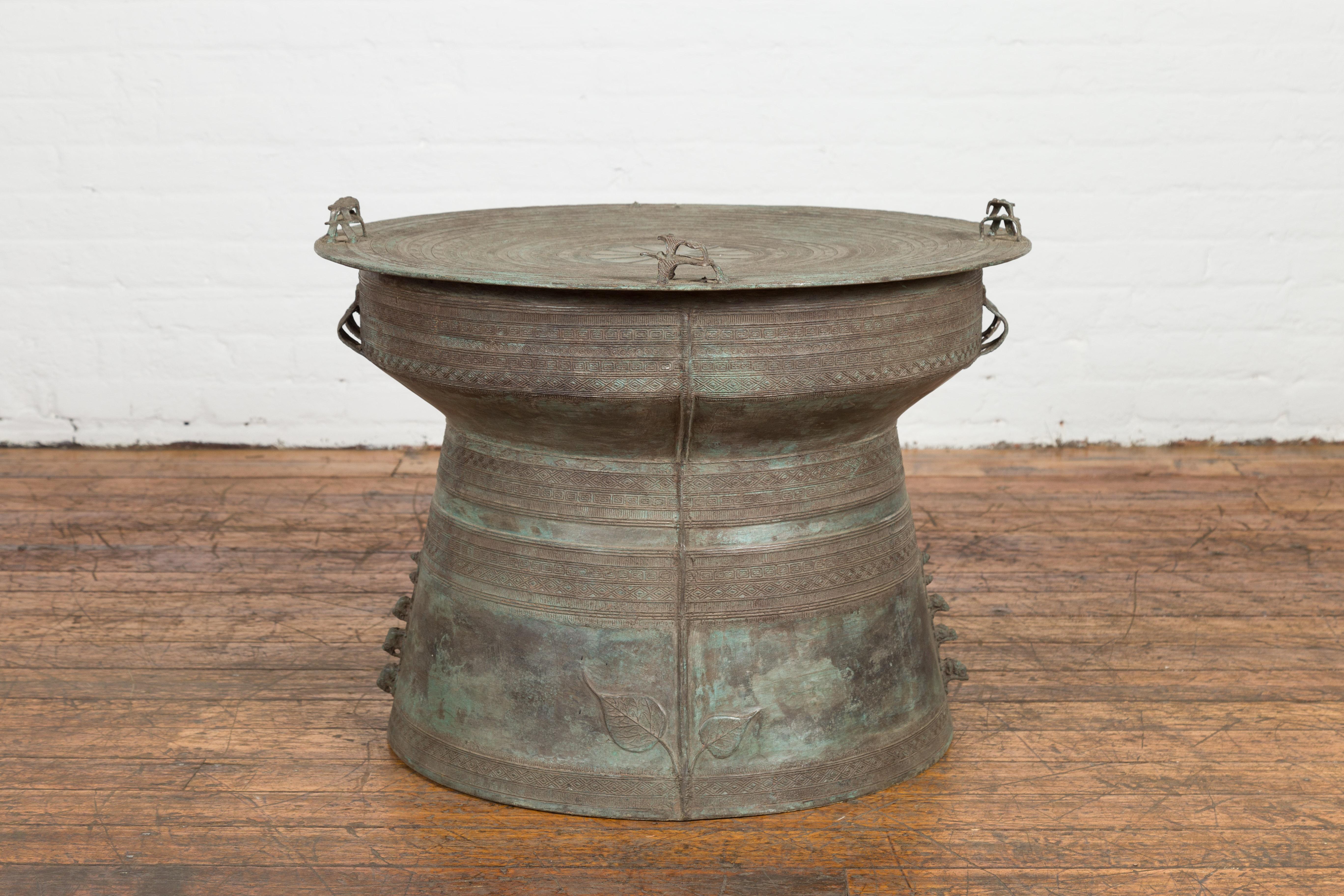 A Laotian style bronze rain drum side table from the Mid-20th Century, with geometric relief motifs, central sun and frog finials. Created during the midcentury period, this bronze drum table (named for the lovely sound that such tables make when