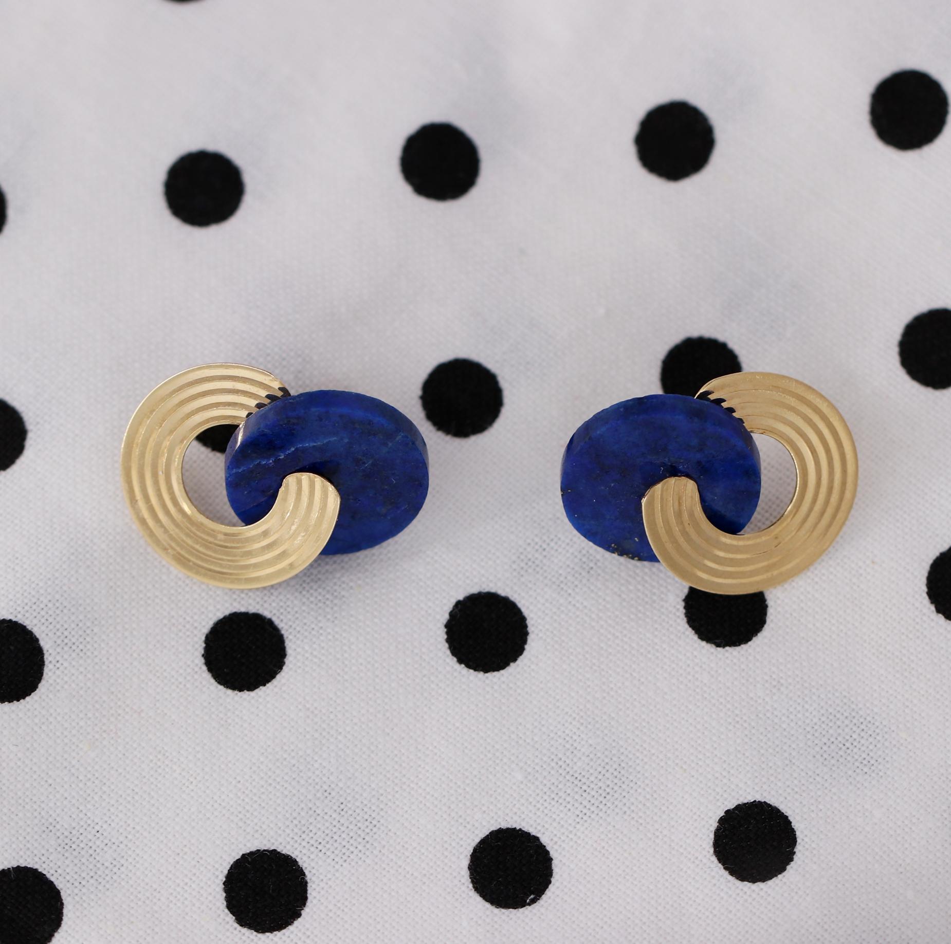 A pair of 14K yellow gold earrings comprised of a 15mm lapis lazuli disc measuring 15mm. A gold post runs through the disc, and is affixed to a grooved piece of gold which creates the appearance of a figure 8 or congruent circles. This modern design