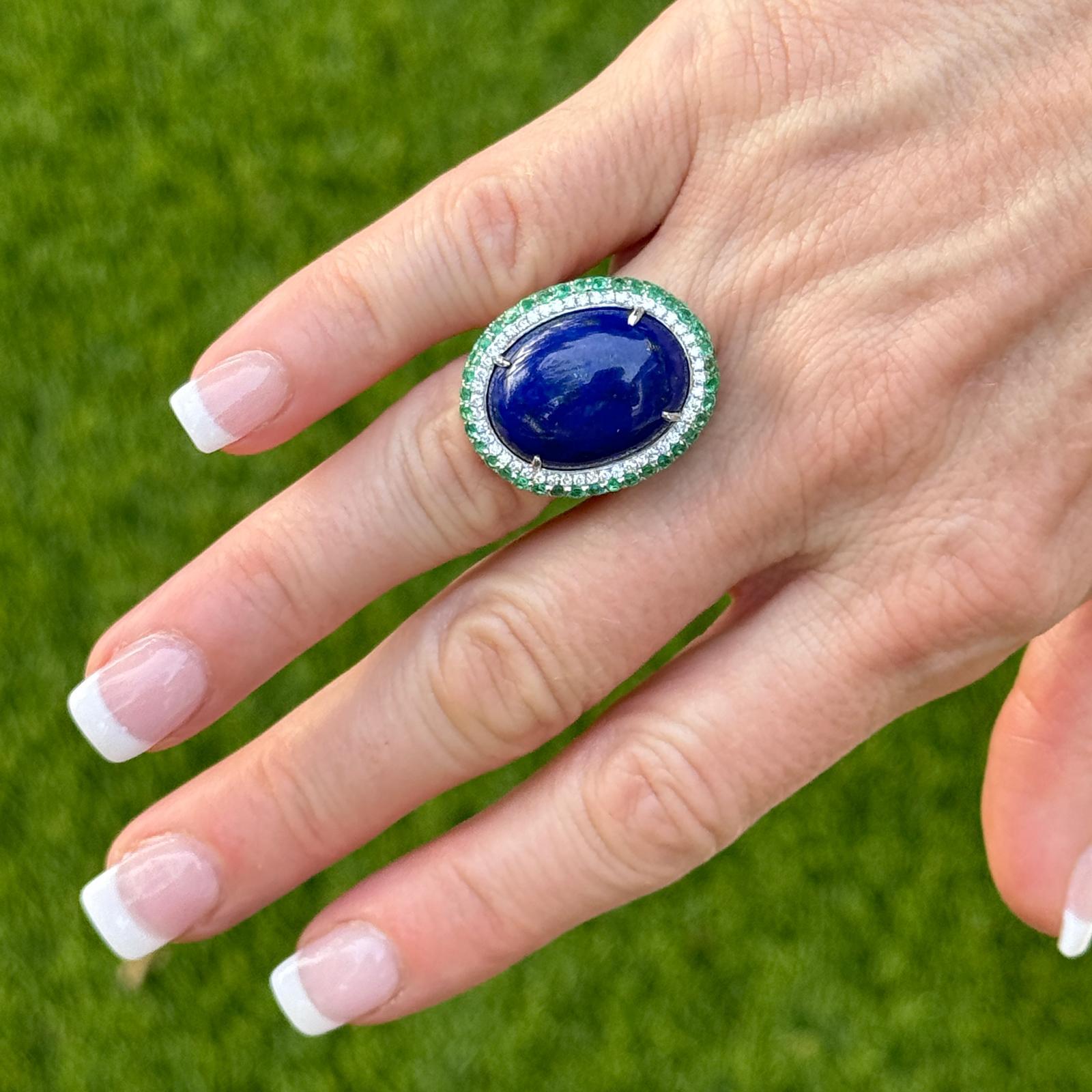 Lapis, emerald, and diamond cocktail ring crafted in 18 karat white gold. The cocktail ring features a cabochon lapis lazuli gemstone, 52 round brilliant cut diamonds, and a row of emeralds. The diamonds weigh approximately .50 CTW and are graded