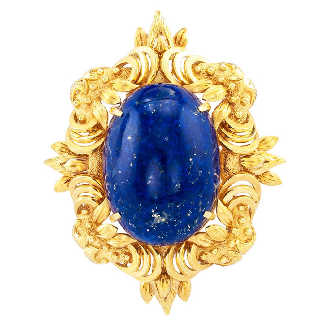 Lapis lazuli and yellow gold brooch pendant. The design showcases a large, oval shaped lapis lazuli cabochon measuring approximately 37 X 27 mm. set in a lavishly decorated gold frame comprising foliar motifs, organic and geometric shapes, to the