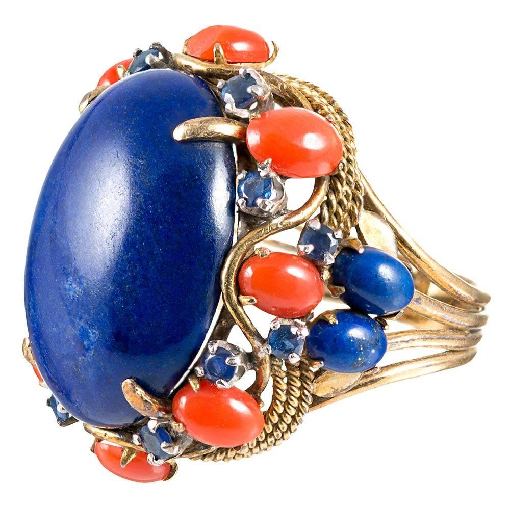 A frame of coral cabochons and blue sapphires dances around a large lapis lazuli centerpiece with a scalloped skirt of golden rope. The ring is boastful in size, yet the absence of diamonds celebrates the gorgeous organic charm of the gemstones