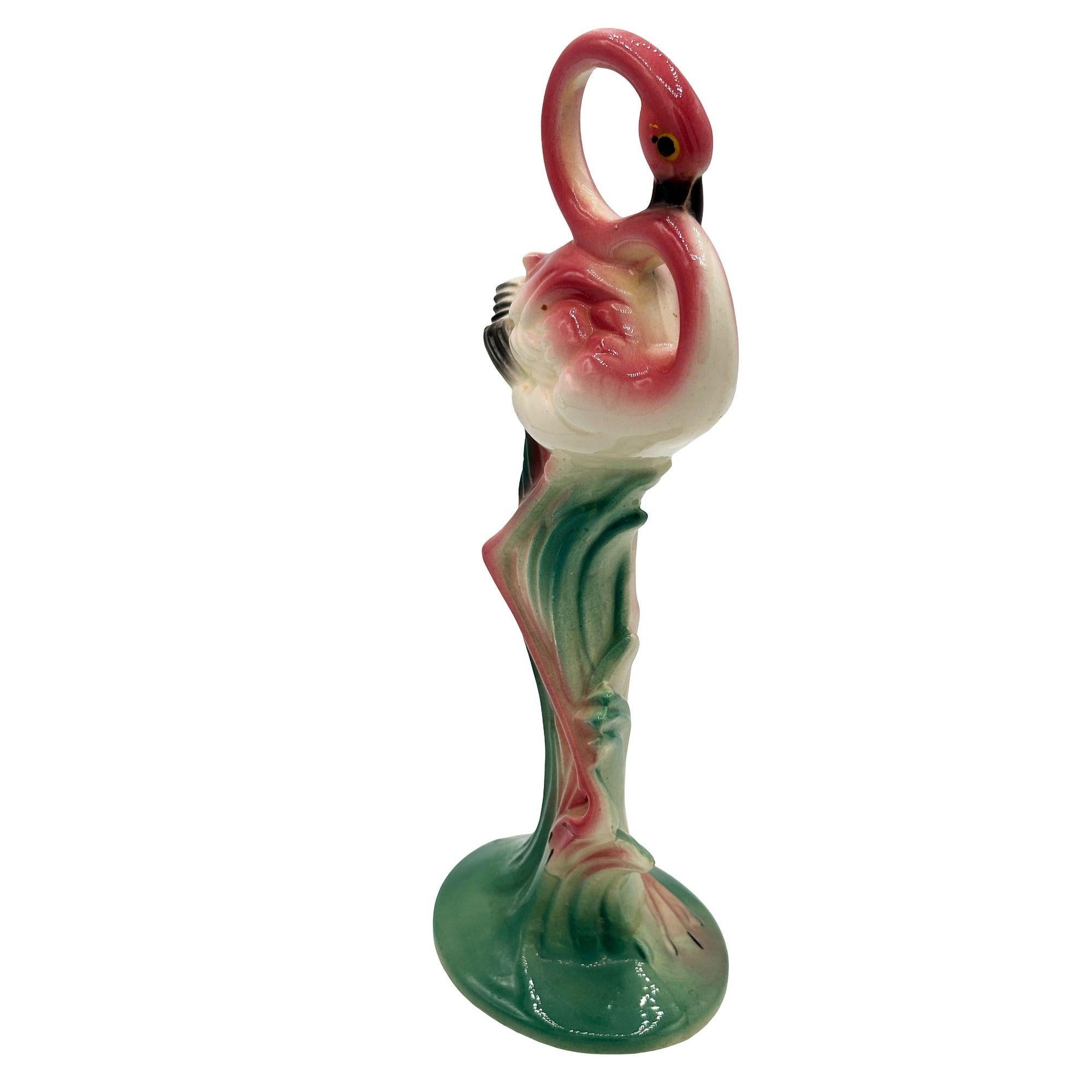 Mid-century pink and green Drinking flamingo ceramic figurine with a bashful expression. The statue detailed a pink flamingo with long legs, a slim, tall form standing on a grassy field, bowing down to take a drink. It comes in a gloss finish that