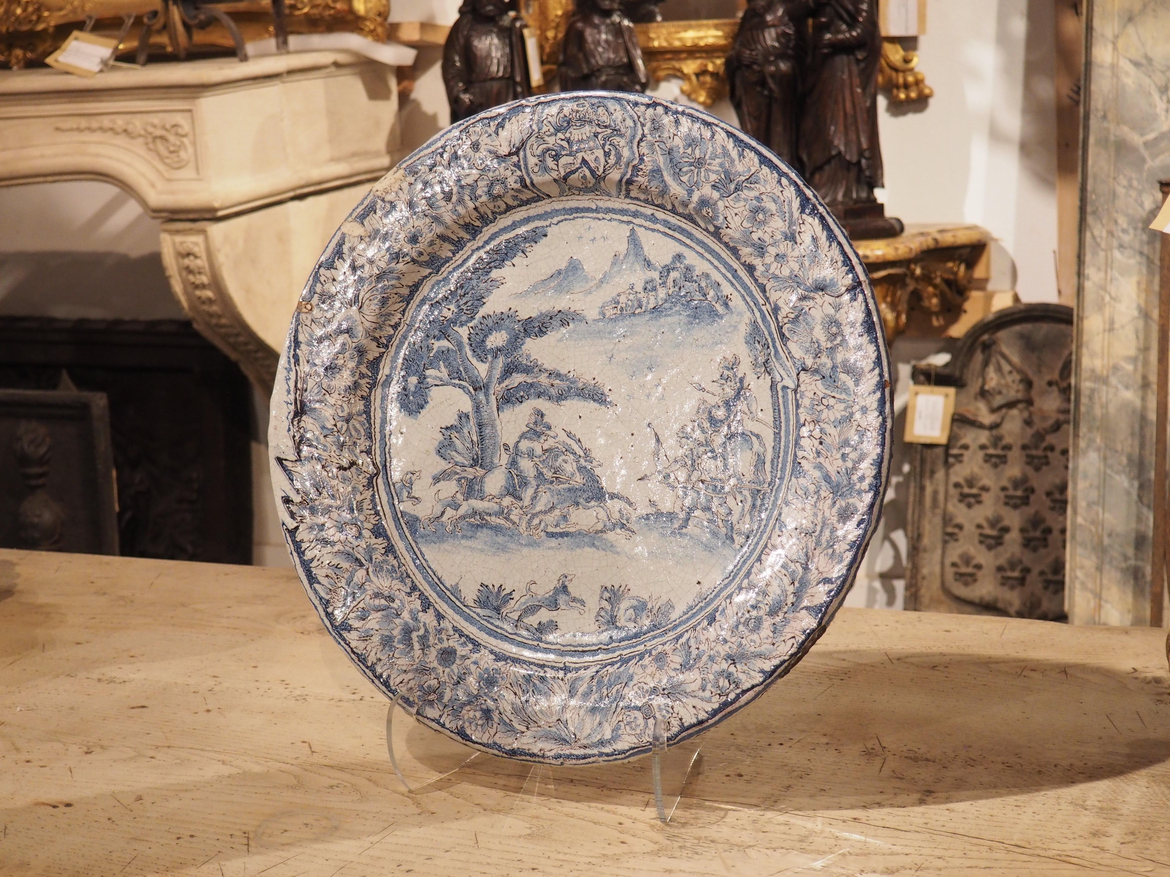 Hand painted in the 1600s in France (most likely in or around Paris), this large blue and white faience platter features a scene depicting a stag hunt surrounded by a luxurious floral wreath on the raised lip. At the top of the platter, set amongst