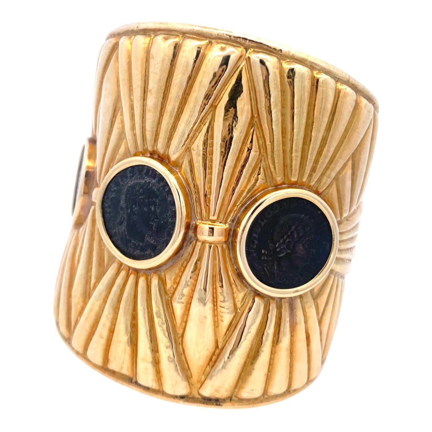 18K yellow gold cuff bracelet, displaying a bold, textured design with three bezel set roman coins. The coins are evenly spaced, creating a striking visual rhythm. The gold surface features radiating lines, enhancing the classical Roman