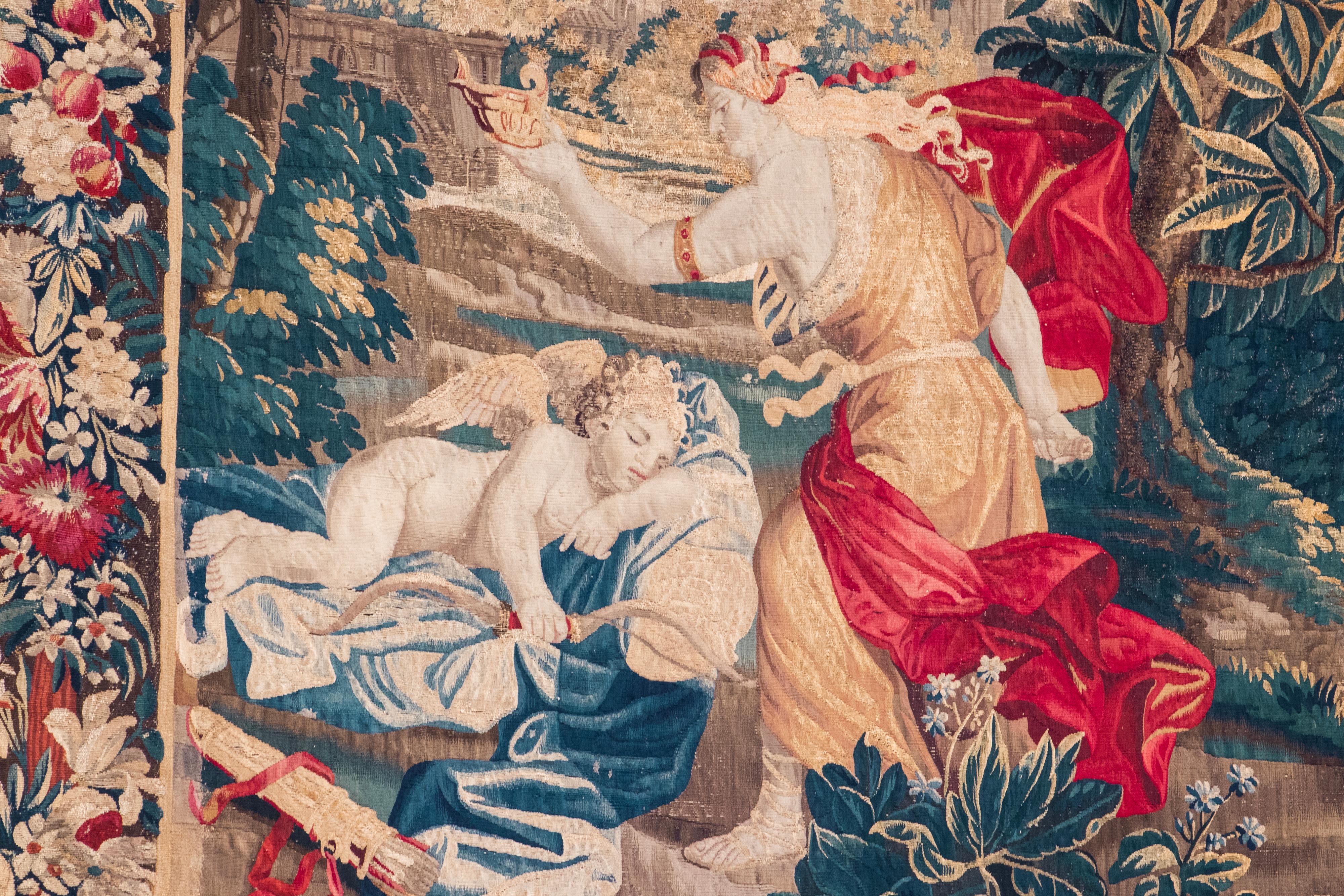 Spectacular Flemish Verdure tapestry depicting Pysche and the Sleeping Cupid in a flourishing garden, with an architectural landscape on the background. The composition is framed with a wide rim, decorated with traditional scroll motifs, florals,