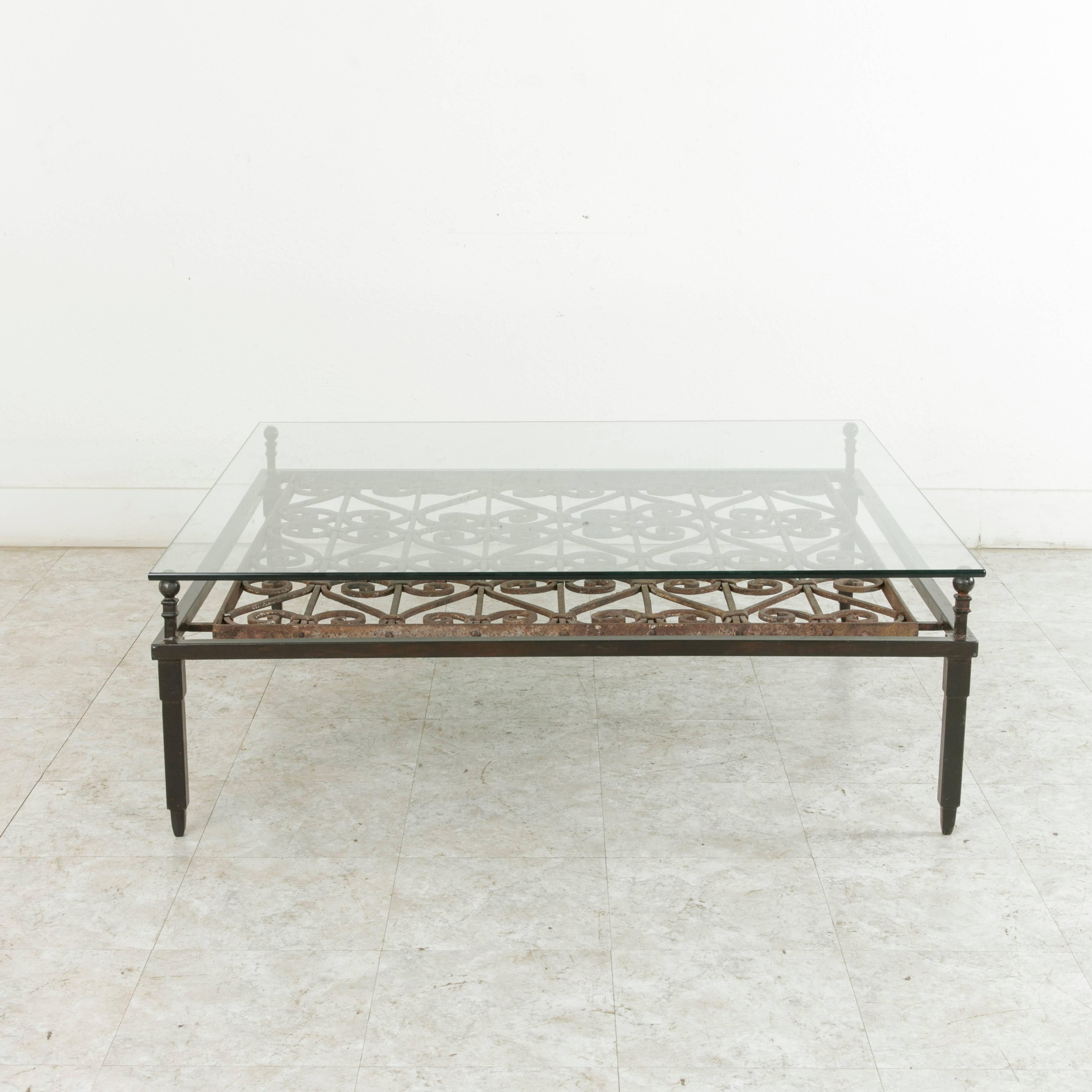 This very large glass top coffee table or garden table measures 48 inches in length and 46 inches in width and features a late eighteenth century hand forged iron grill. The glass top, mounted on the four legs rising above the iron frame, highlights