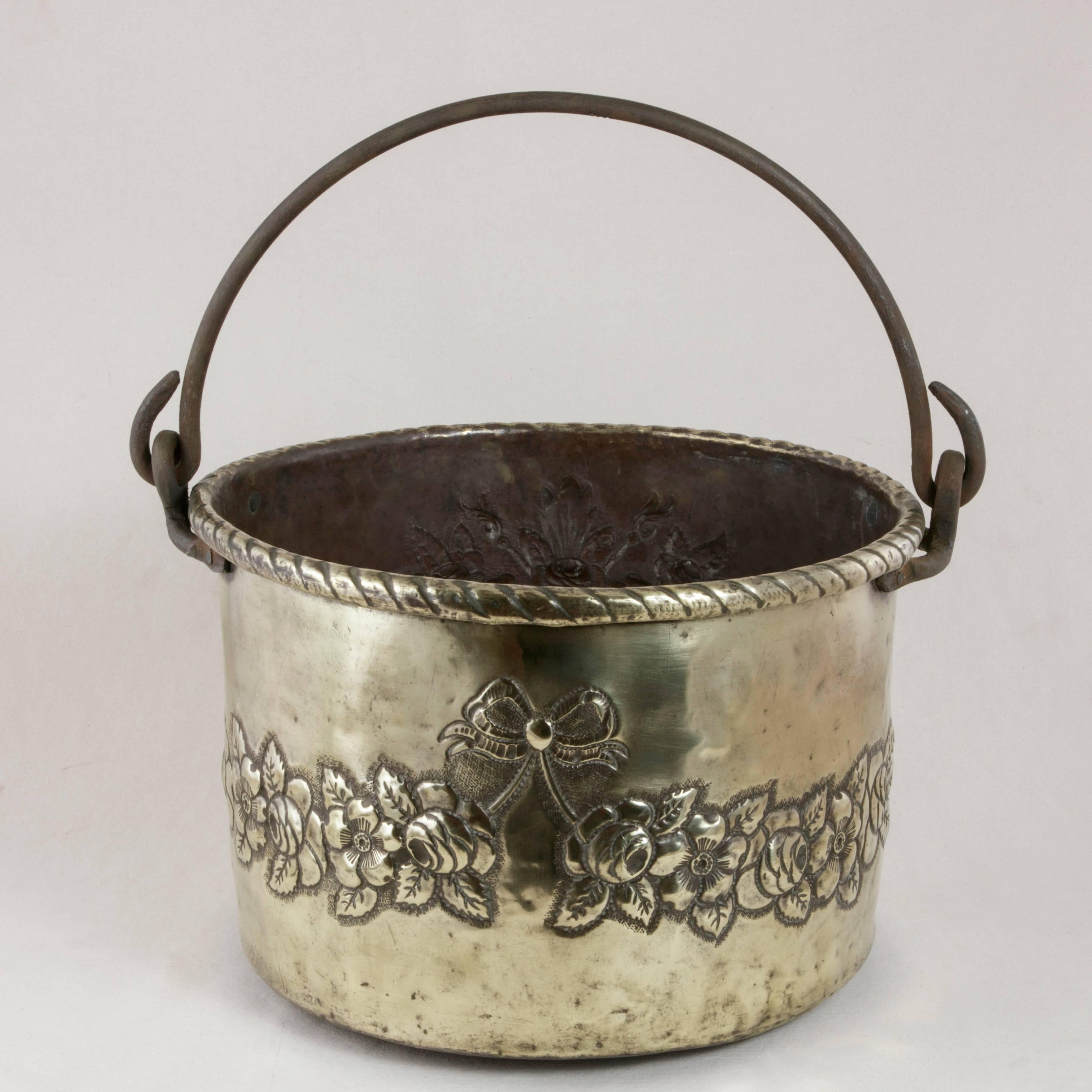 This late 18th century French brass repousse cauldron features a basket and floral bouquet on one side and a garland of flowers with a central ribbon and bow on the other. A twisted ribbon motif surrounds its upper rolled edge. This large pot is