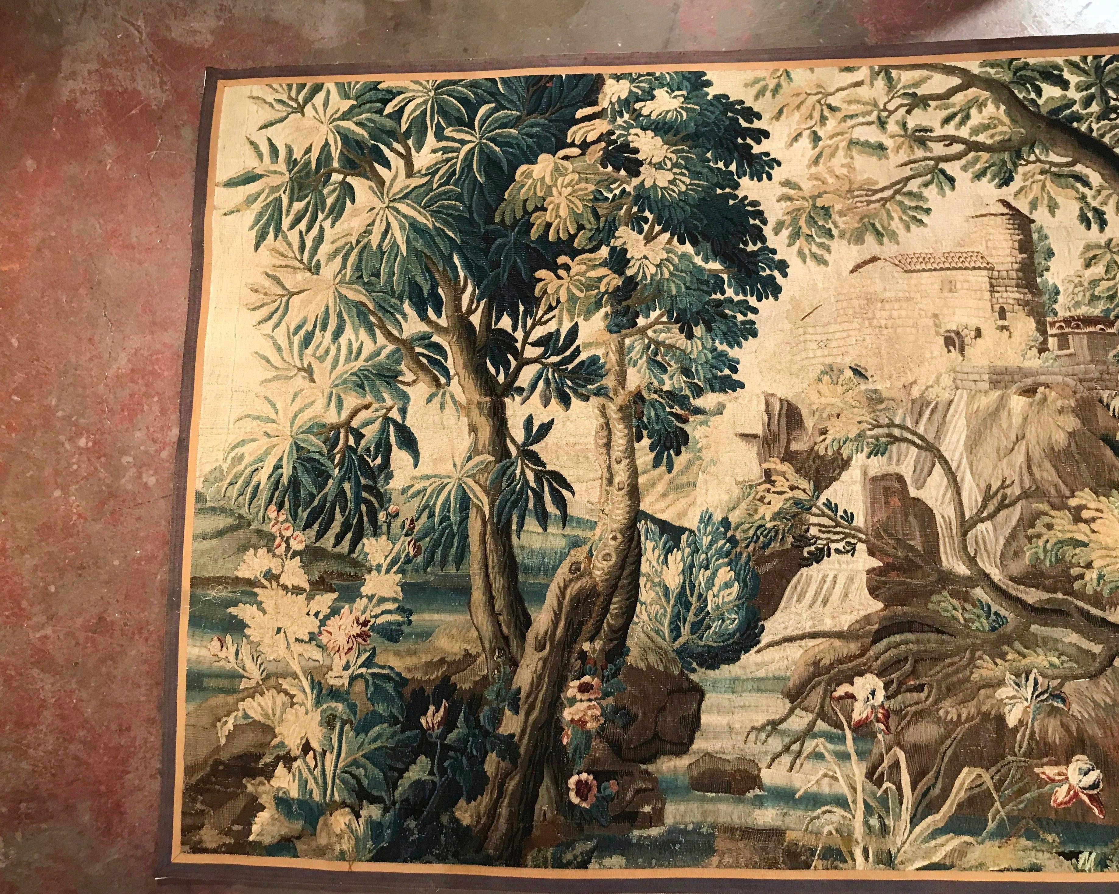 This colorful antique tapestry was found in Versailles, France; handwoven in the village of Aubusson, the wall hanging piece features foliage decor with a running stream, an old structure in the background and a large bird standing on a fence. The
