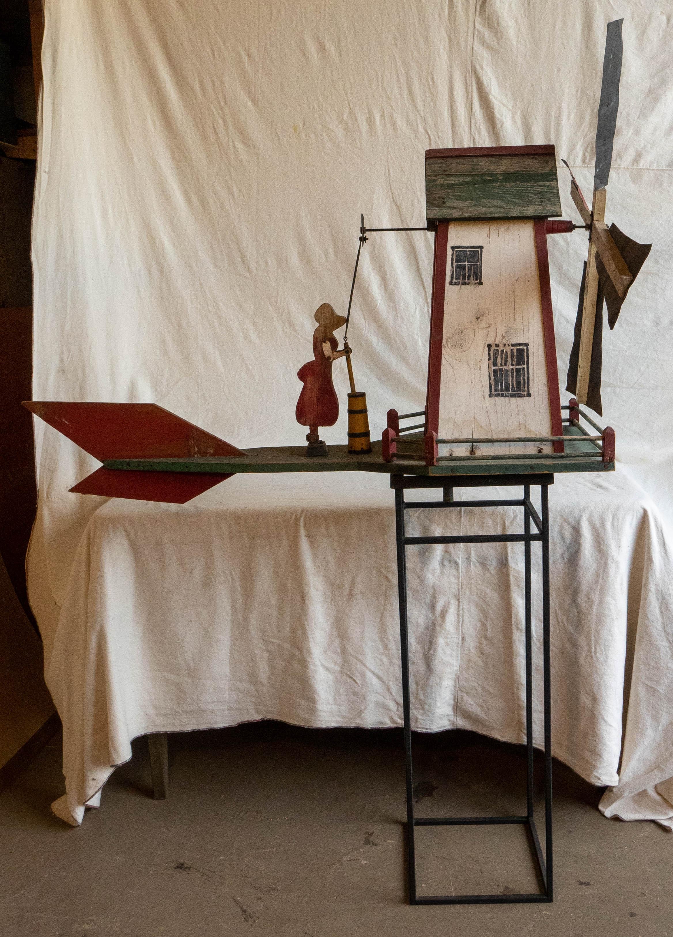 Large scale windmill whirligig with woman churning butter. In very good condition with vibrant red, green, white, and yellow painted details. Sits on iron stand. Height with stand 70.5 inches.