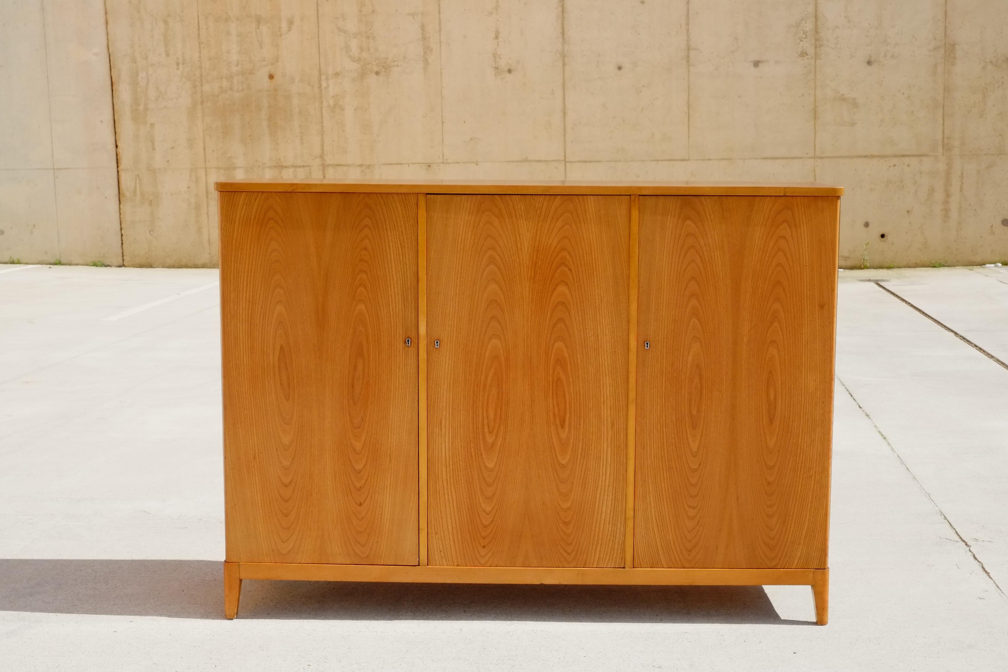 A beautiful 1950s Art Deco sideboard by Carlsson & Reicke, Stockholm. This sideboard has three tall doors in a beautiful honey coloured, book matched elm wood. The interior is a lovely contrasting pale birch wood with two shelved areas to the left
