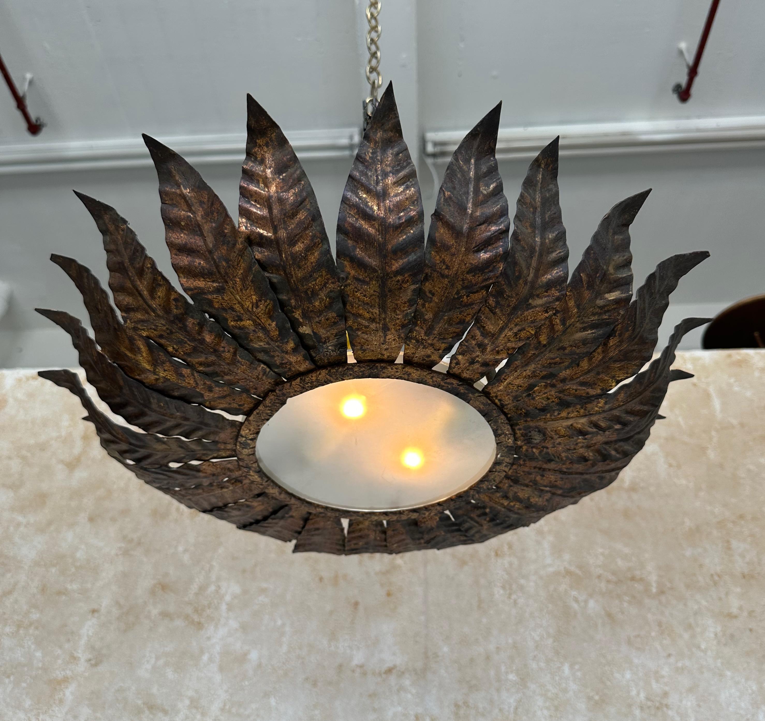 Ce plafonnier espagnol des années 1950, fabriqué à la main, présente une couronne de grandes feuilles qui s'incurvent gracieusement vers le haut et entourent un centre circulaire en verre dépoli. La patine dorée richement appliquée à la main est