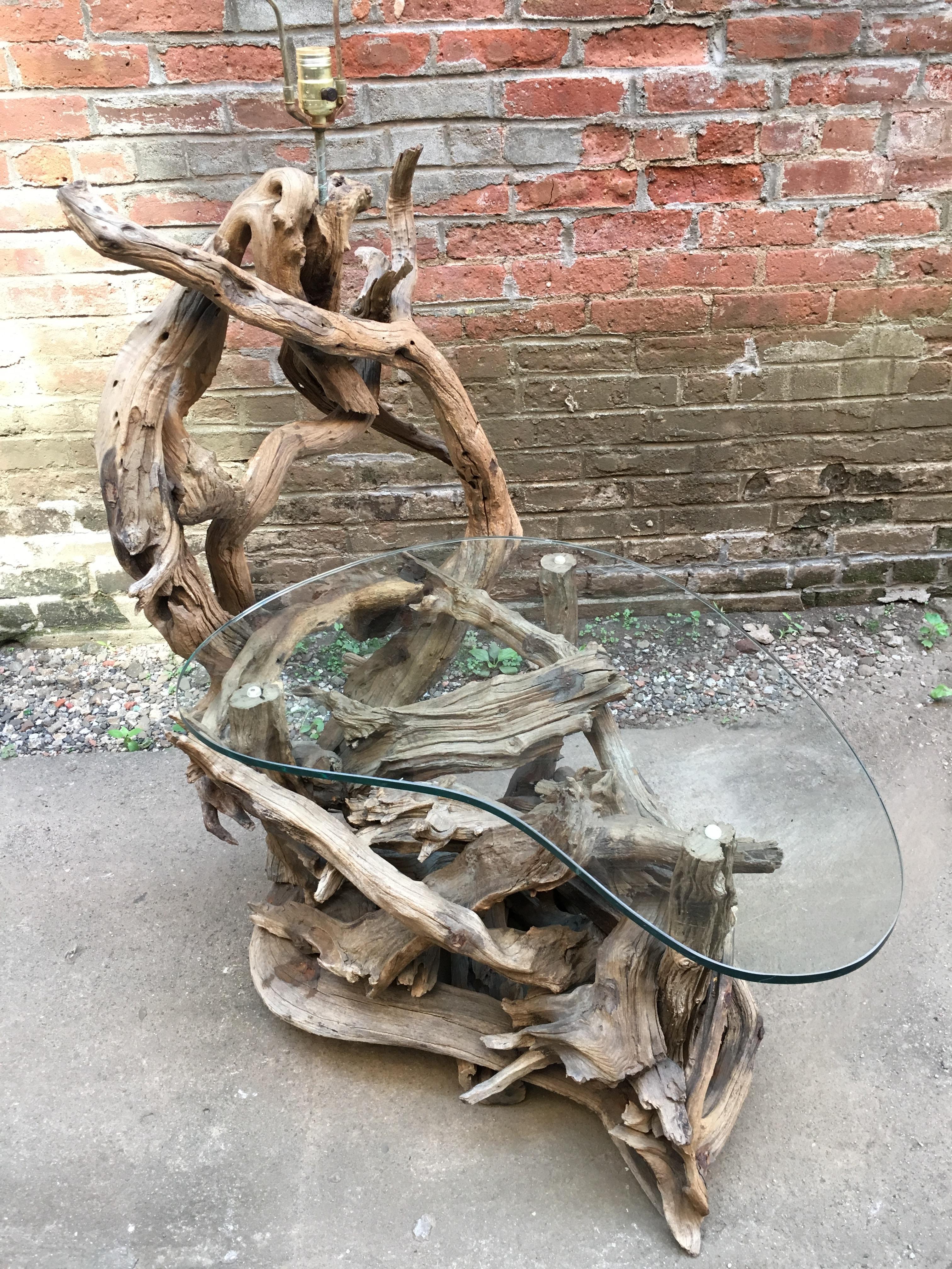 A large twisted, sculptural mass of driftwood that has been artfully rendered into a floor lamp and a table, circa 1960. The base holds a boomerang shaped piece of glass. Weathered to perfection.