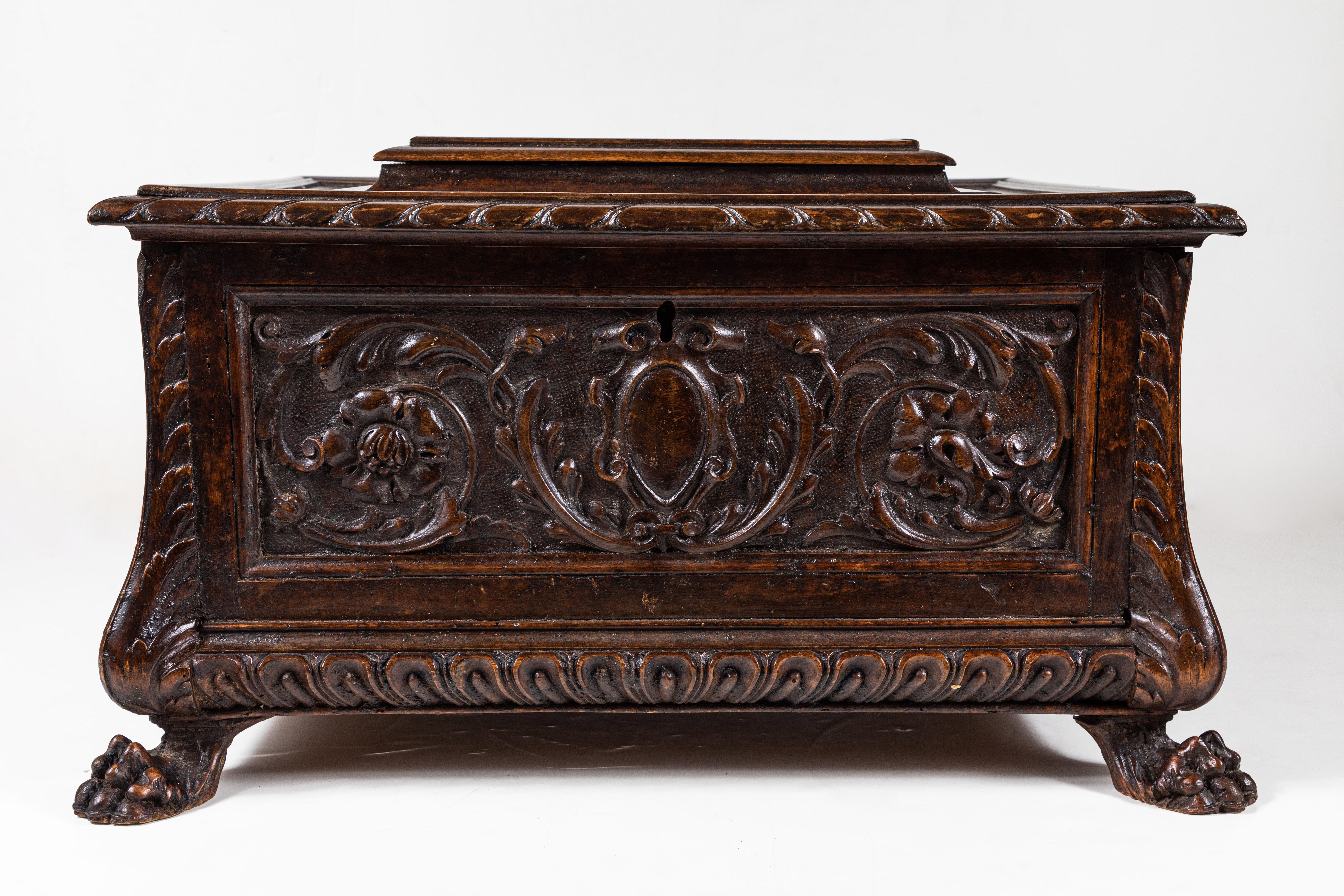 Grand, lidded, hinge-top, richly carved walnut coffer embellished with four panels of reliefs featuring shields bordered by foliate scrolls. The whole atop paw feet.