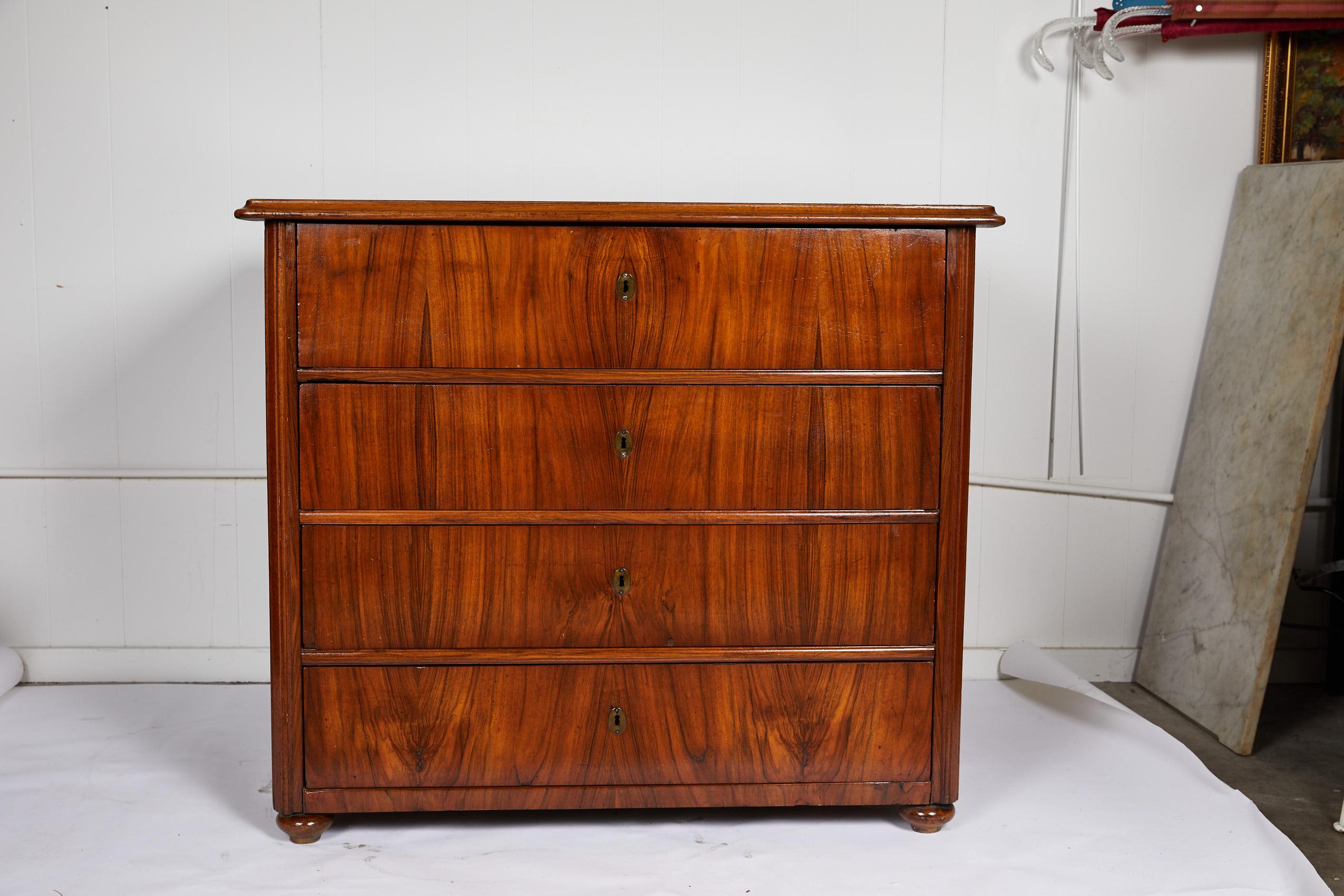 19th century large scaled and sleek Continental Biedermeier commode with a burl bookplate rosewood veneer on three sides. The case has a rectangular one board top and holds four deep drawers with oval brass escutcheons. The commode rests on bun feet.