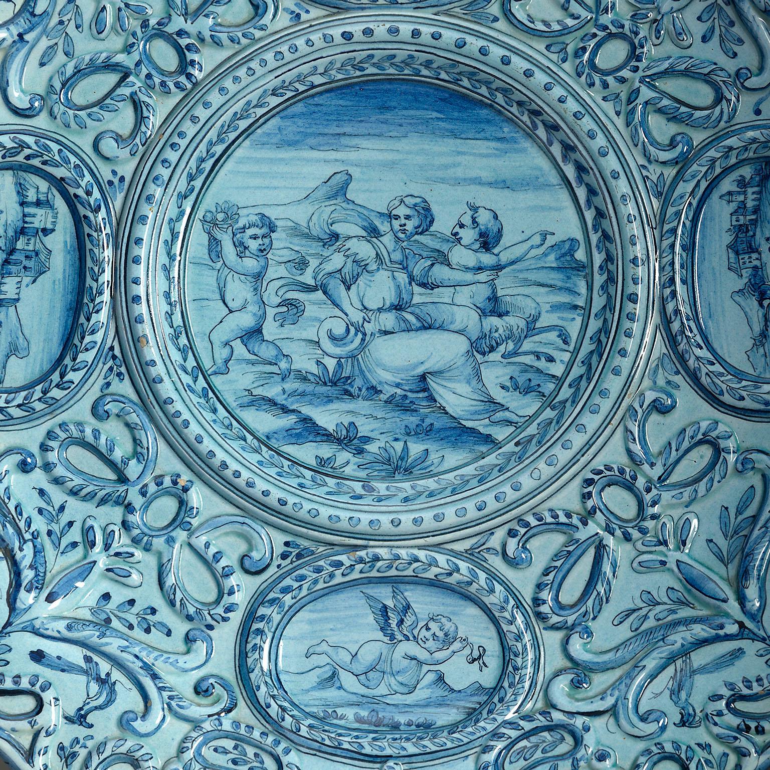 A late nineteenth century blue and white glazed Maiolica charger of large scale, decorated with floral and foliate work, the central circular roundel and surrounding oval cartouches with cherubs and landscapes.

Purchased from a private collection